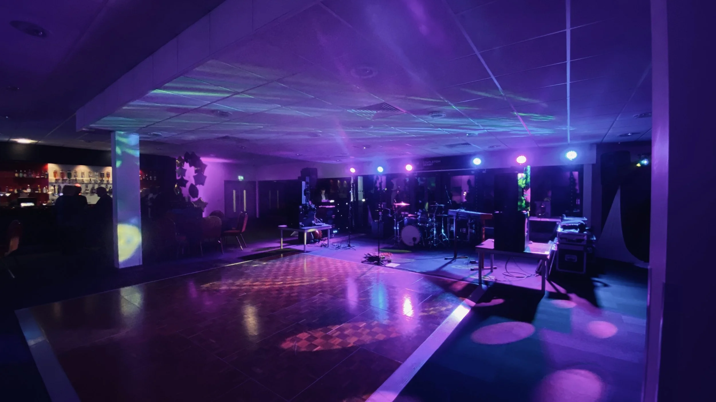 Empty dance floor with stage for live music, illuminated by colorful stage lights in purple, blue, and pink in a nightclub or event space