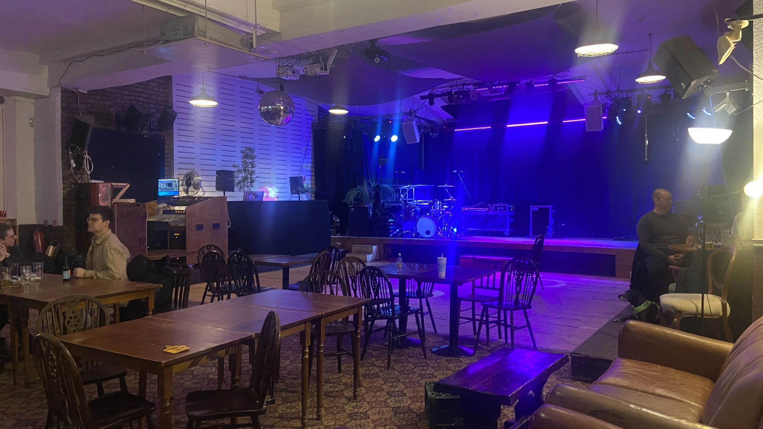 An empty stage with musical instruments and purple stage lighting in a dimly lit bar or club. Seating area with wooden tables, chairs, and a leather couch, and some people sitting and standing around.