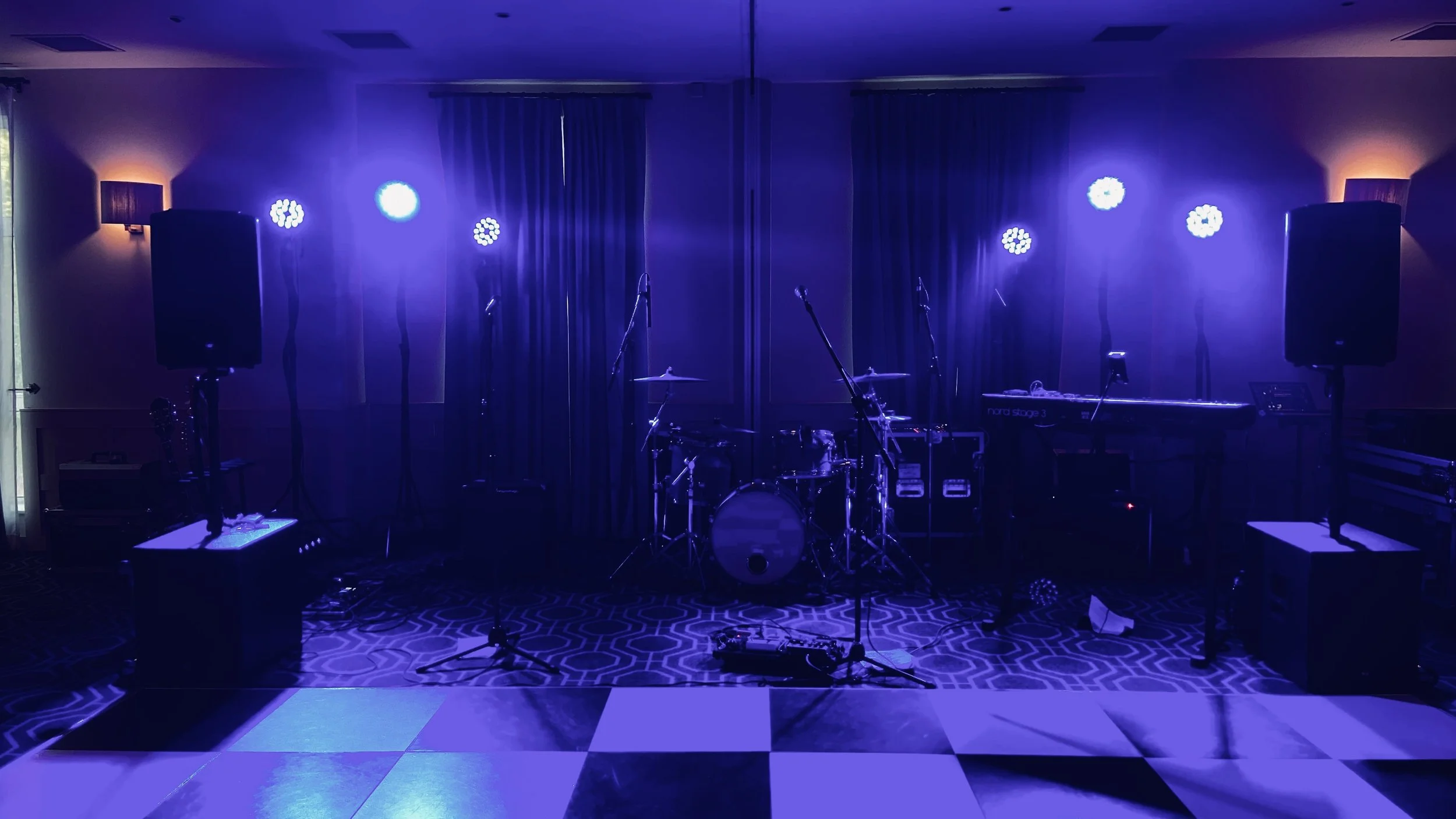 Stage setup with drum kit, keyboard, microphones, and speakers, illuminated by blue stage lights in a dimly lit room.