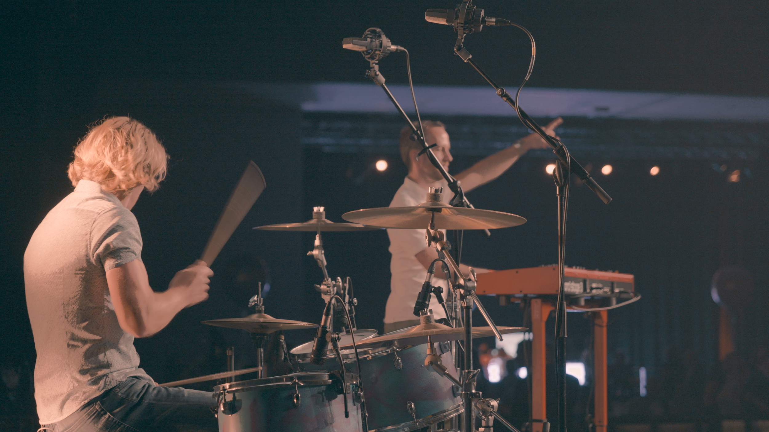 A person playing drums and another person standing in front of microphones on a stage.