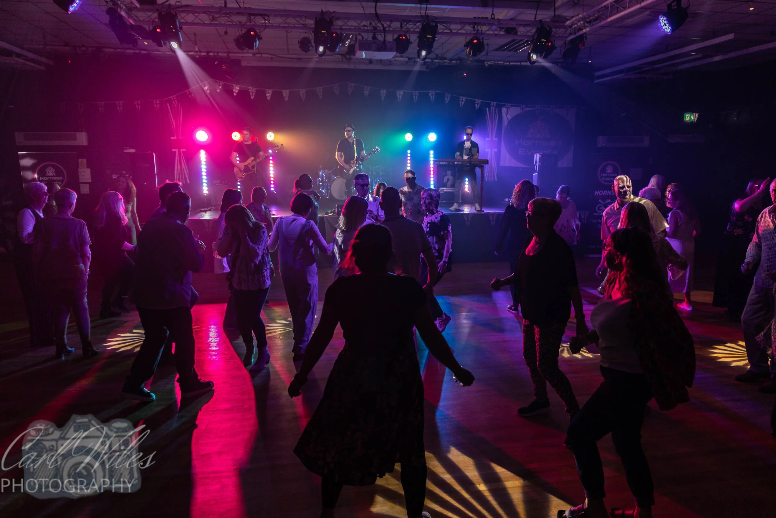 People dancing on a floor in front of a stage with live band playing, colorful stage lights, and some with painted faces at a party or concert event.