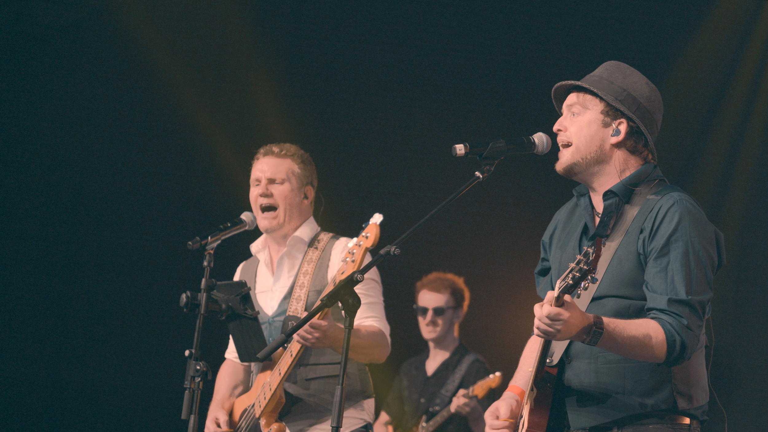 Musicians performing on stage, singing and playing guitars, with one wearing sunglasses and another with a hat, under stage lighting.