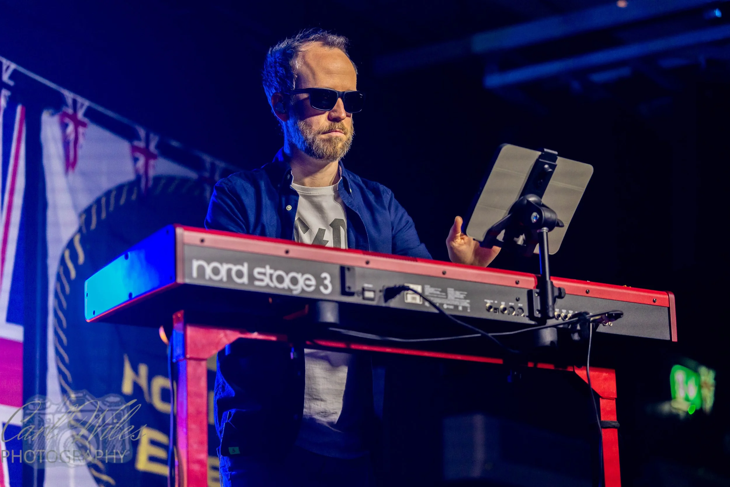 A man with a beard wearing sunglasses and a blue shirt playing a Nord Stage 3 keyboard on stage with a dark background and stage lights.