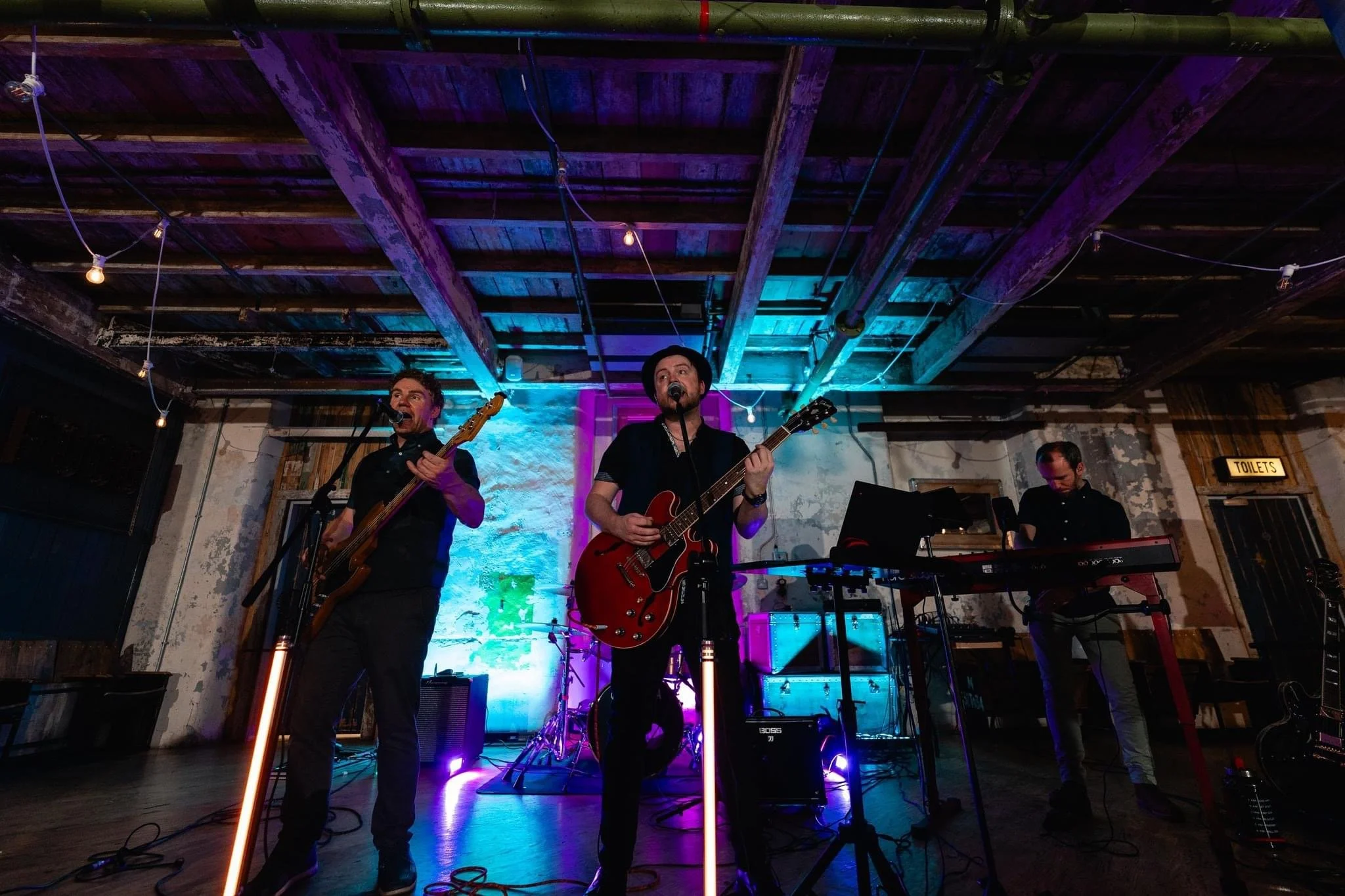 Band performing on stage with three musicians, two singing into microphones, one playing keyboard, with colorful lighting and an exposed wooden ceiling.