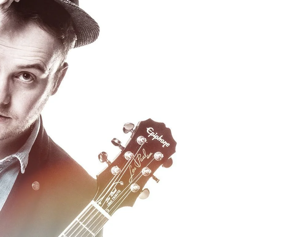 A man wearing a hat and a shirt, partially visible on the left side, holding an Epiphone Les Paul electric guitar against a plain white background.