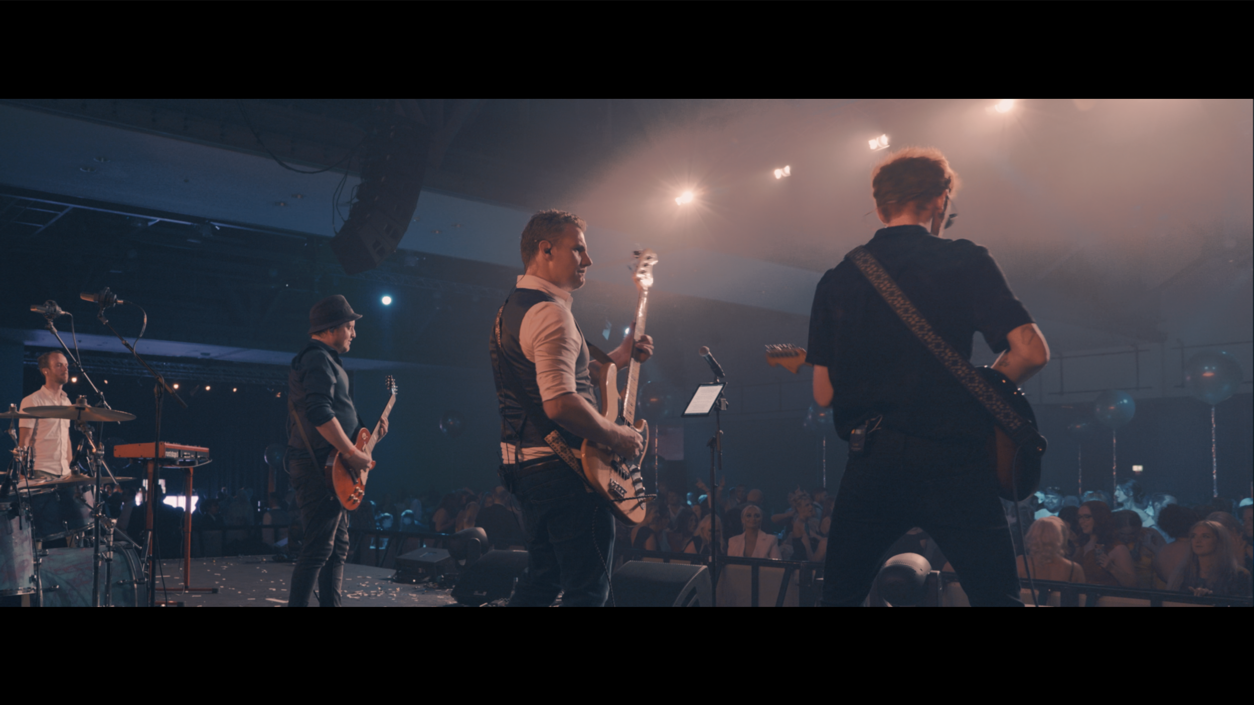 A band performing live on stage with four musicians playing guitars and drums, facing an audience, in a concert hall with stage lights and decorations.