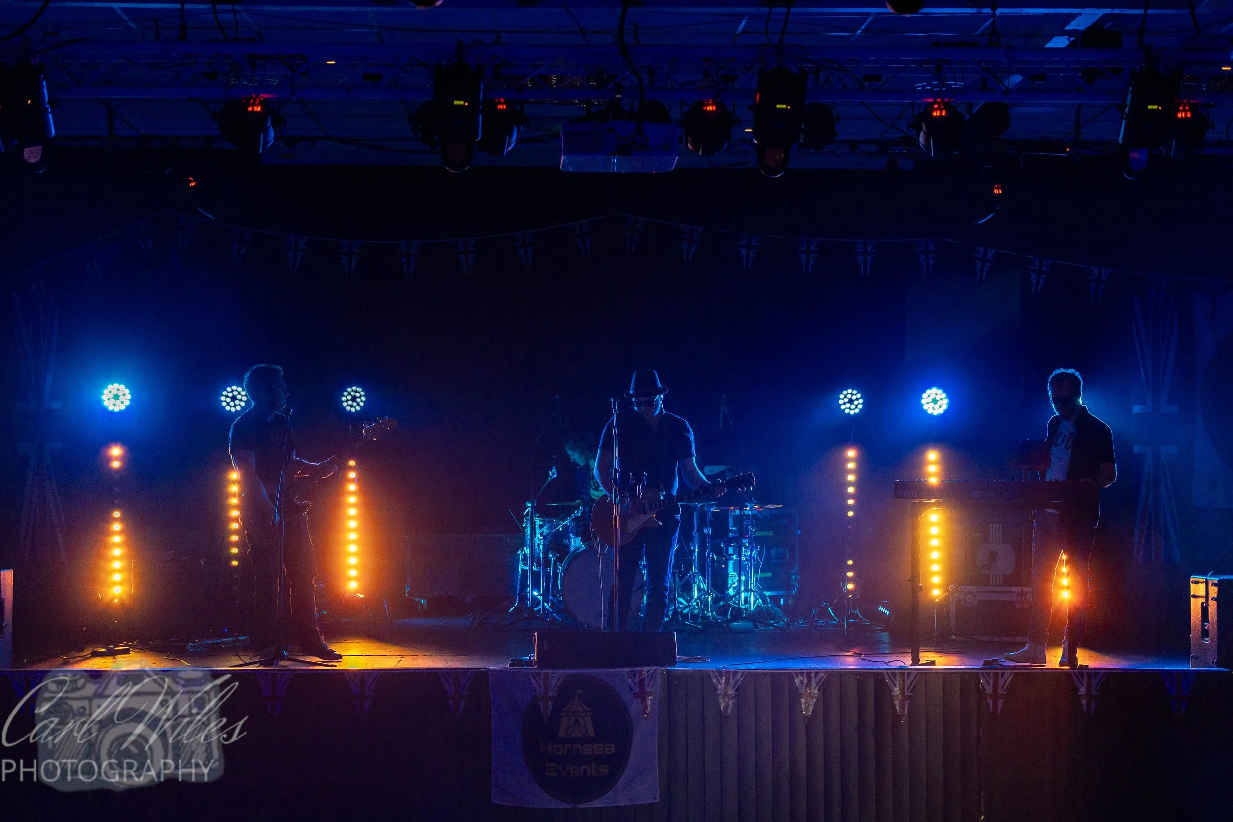 A band of four musicians performs on stage under blue and yellow stage lights, with one musician playing guitar and a drummer in the background, in a dark setting.