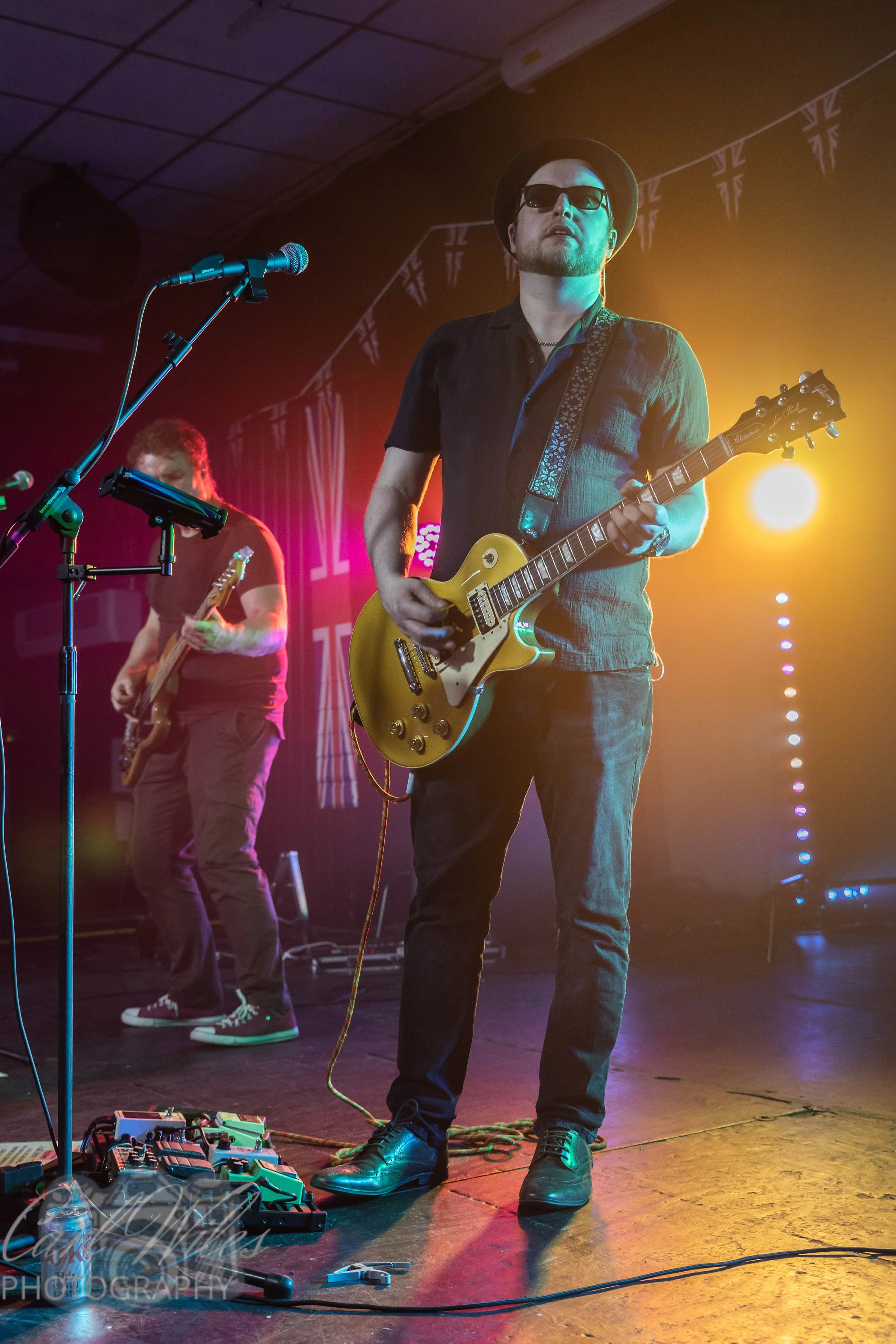 A musician playing an electric guitar on stage with colorful lighting, another guitarist in the background, and various equipment on the floor.