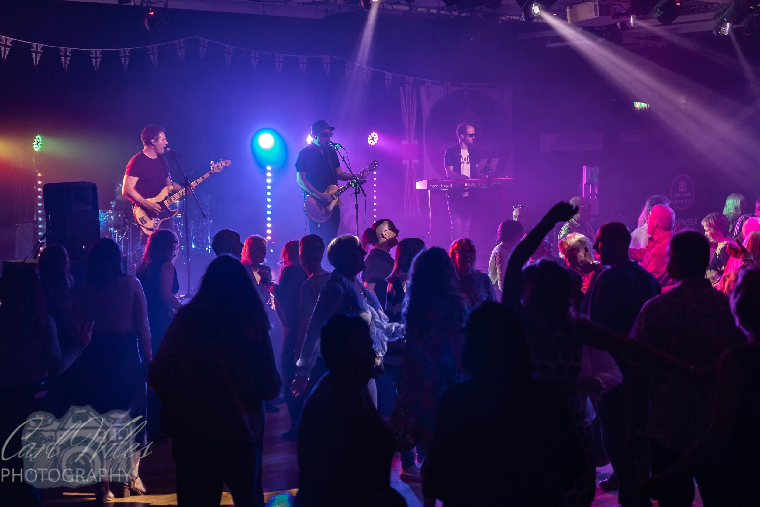 A band performing on stage with colorful lights, with an audience dancing and enjoying the music in a lively indoor venue.