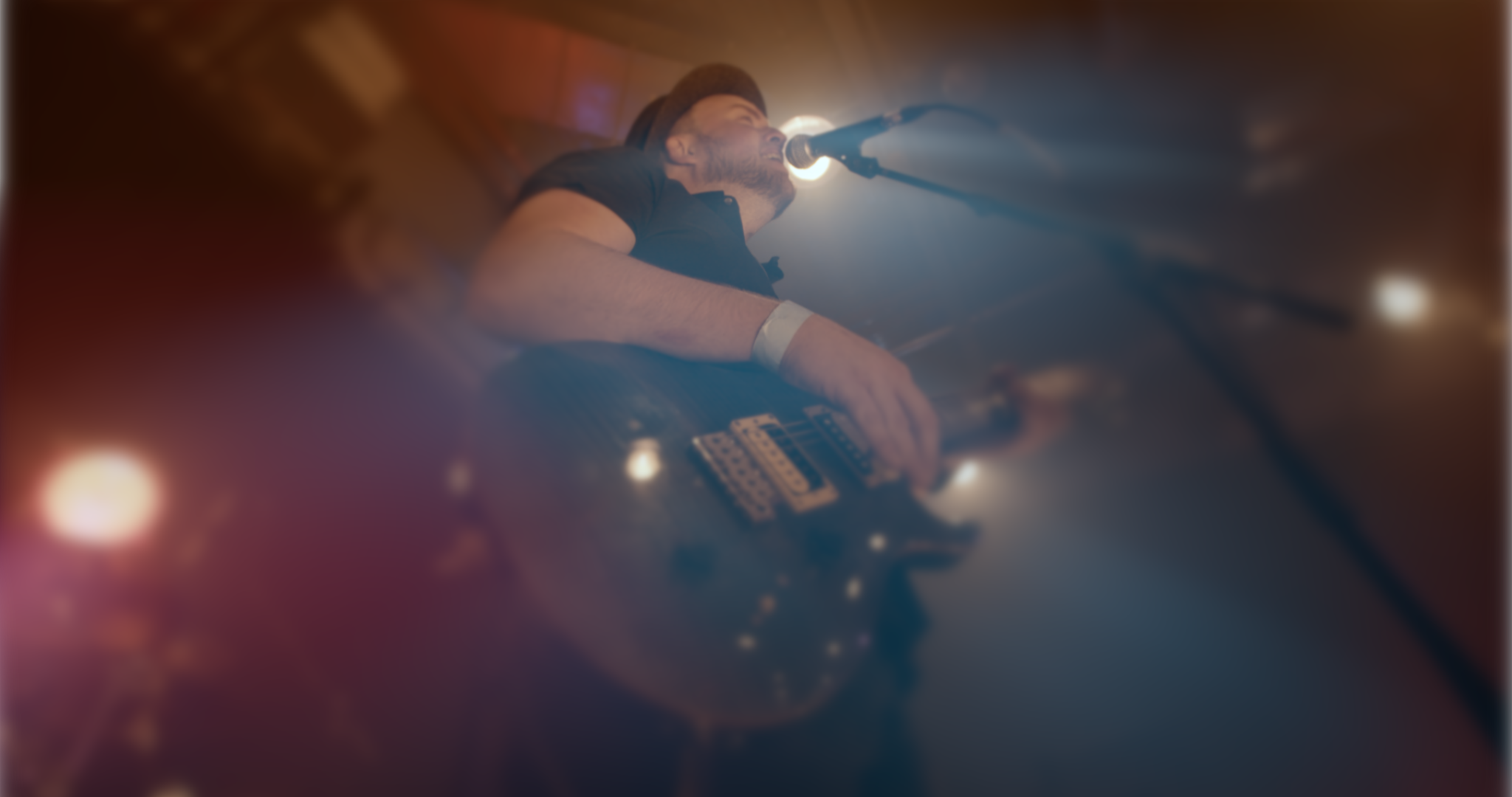 A male musician with a hat playing an electric guitar and singing into a microphone during a live performance in a dimly lit setting.