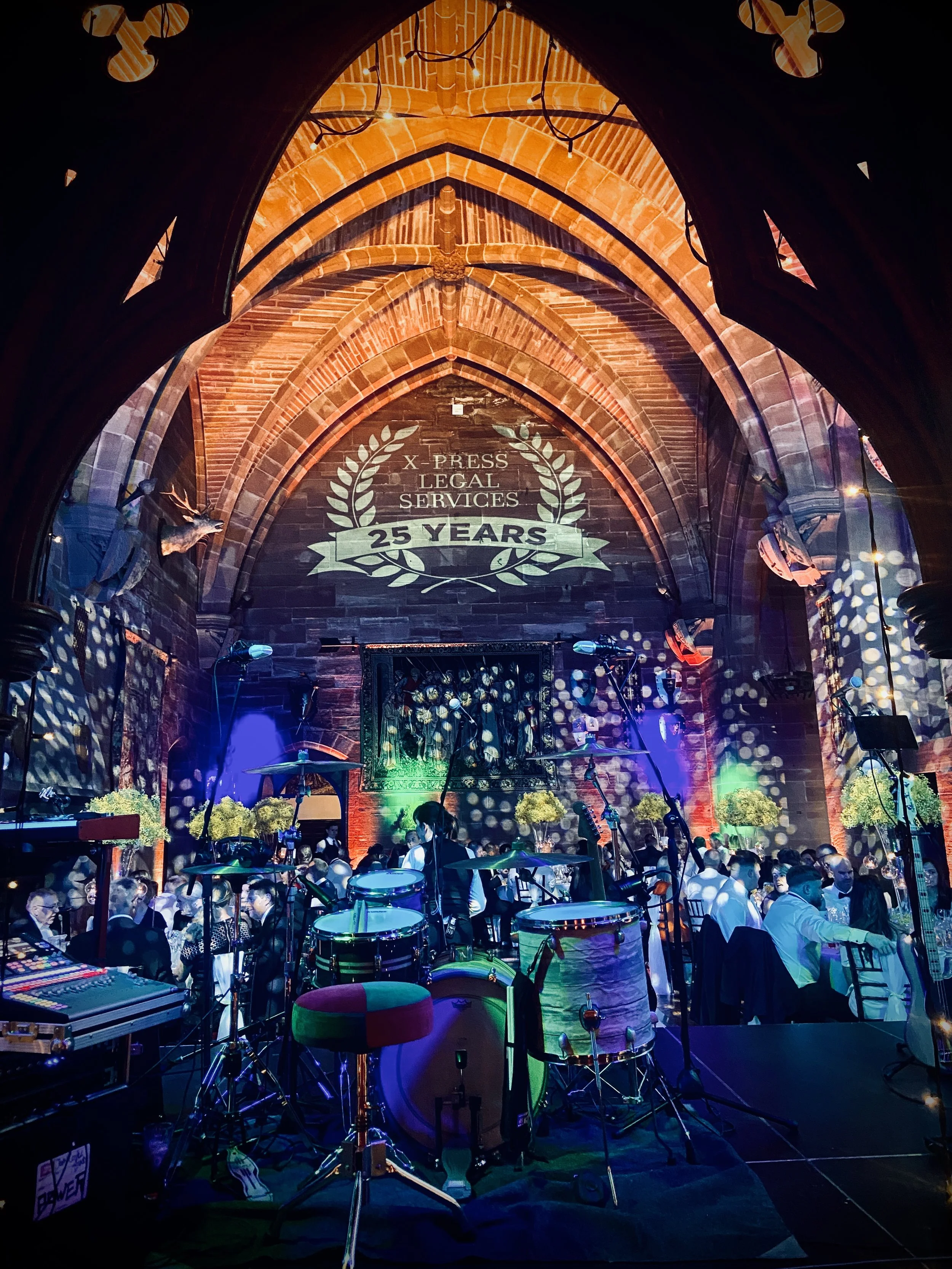 An after-party event celebrating 25 years of X-Press Legal Services in a large, decorated hall with an arched brick ceiling. The room is filled with tables and people dressed in formal attire. A stage with musical instruments, including drums and mic