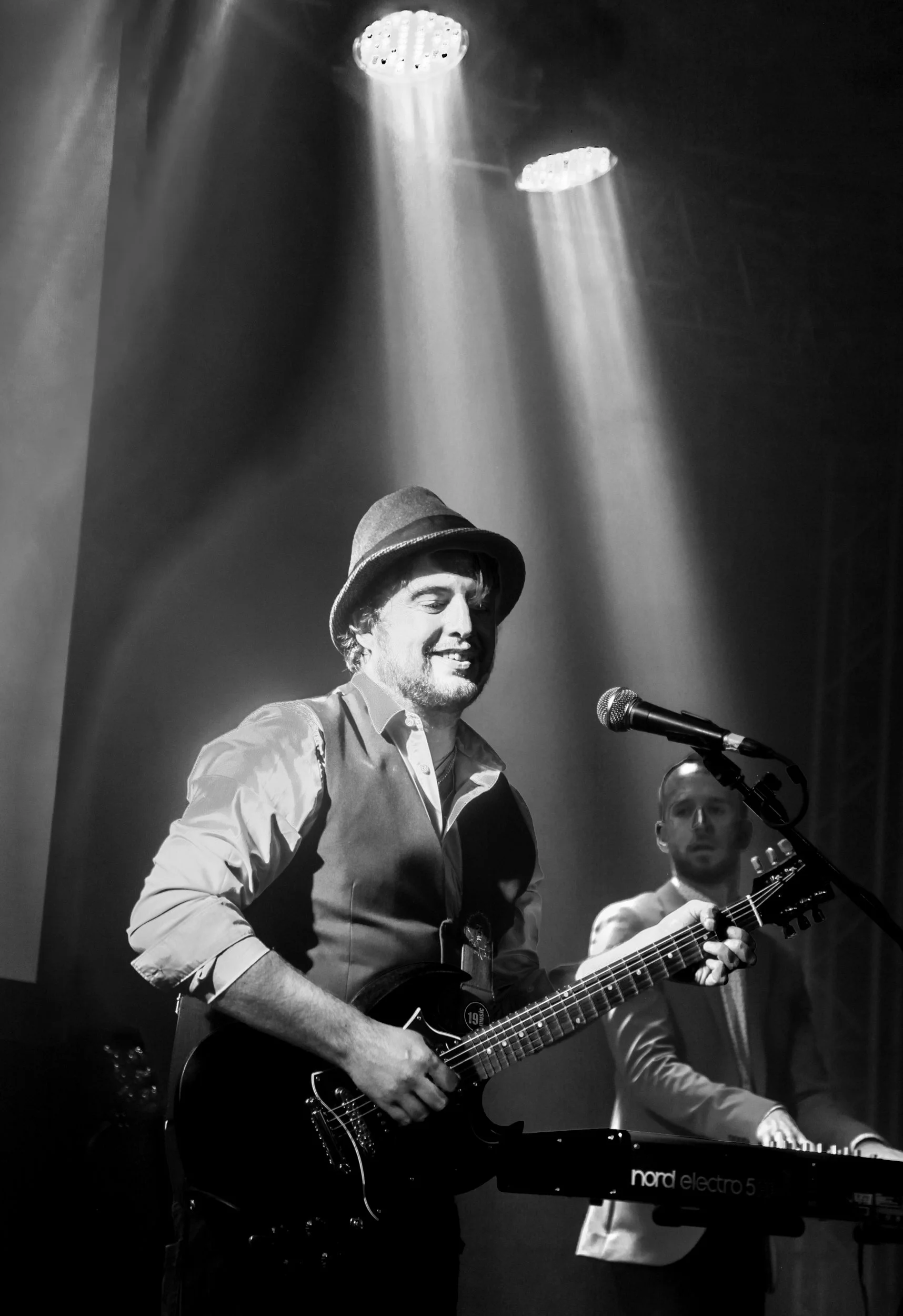 A musician wearing a hat plays an electric guitar on stage under spotlight, with another musician at a keyboard in the background.