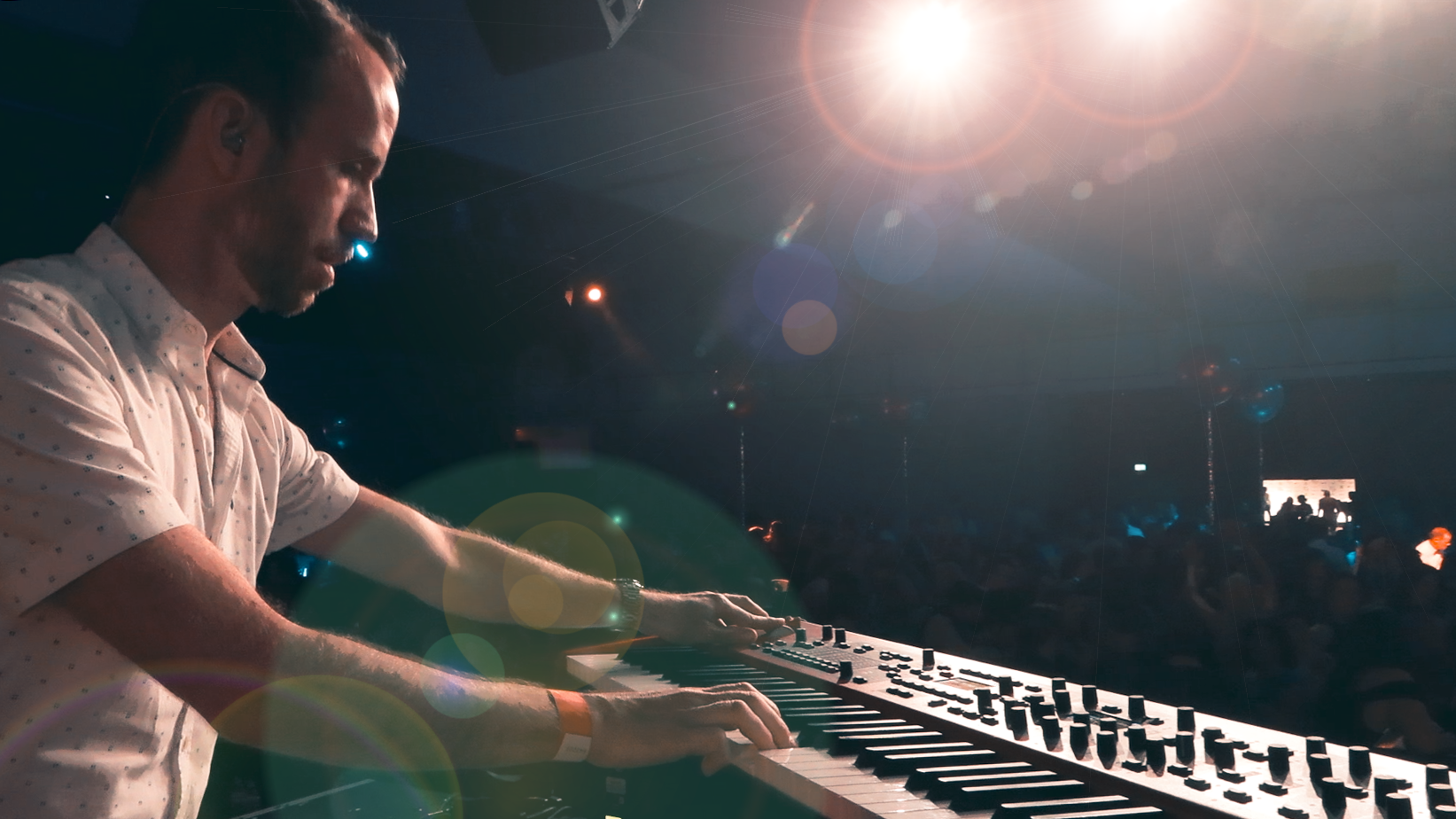 A man playing an electronic keyboard on stage during a live music event with a large crowd in the background and bright stage lights overhead.