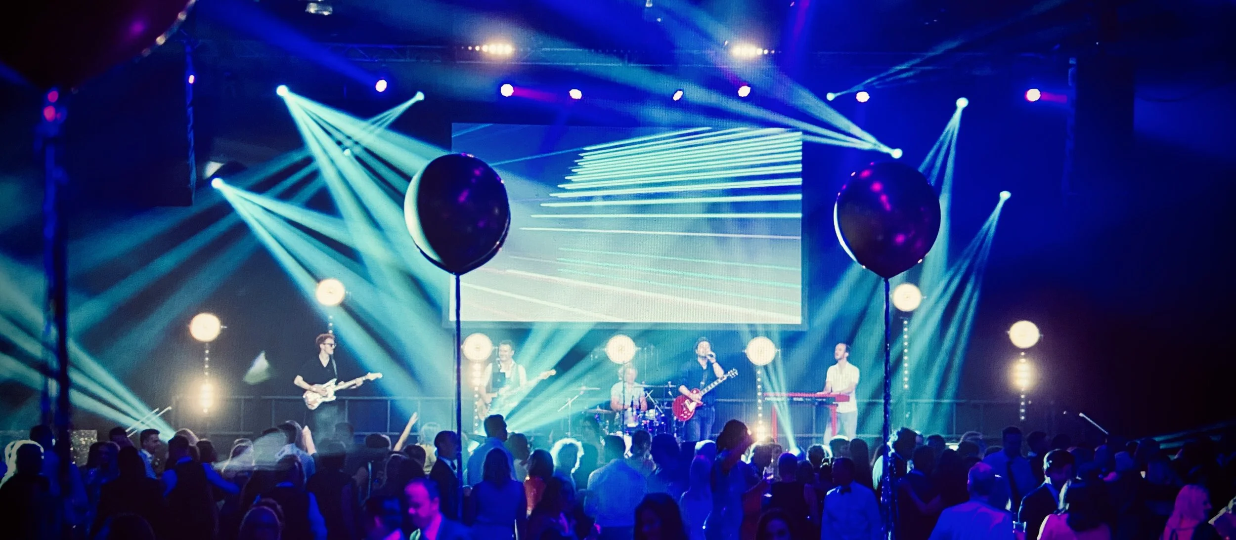 A band performing on stage with colorful lighting and a large screen in the background, while an audience watches in front.