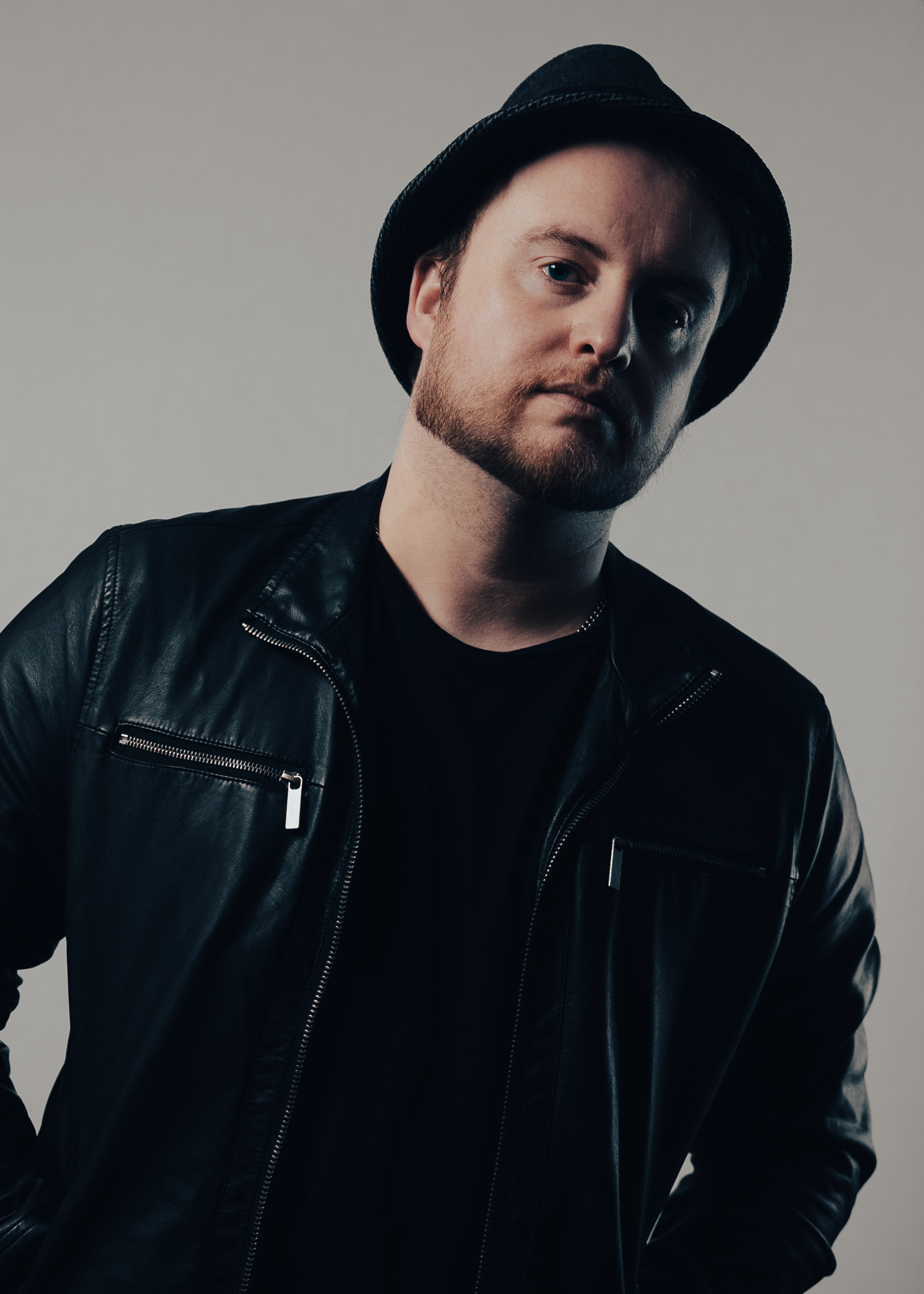 A young man with a beard wearing a black fedora hat, black leather jacket, and black shirt, posing against a plain gray background.