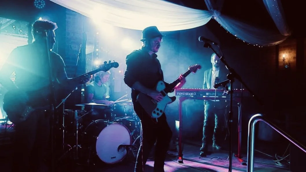 Band performing live on stage with guitars, keyboards, and drums, illuminated by colorful stage lighting