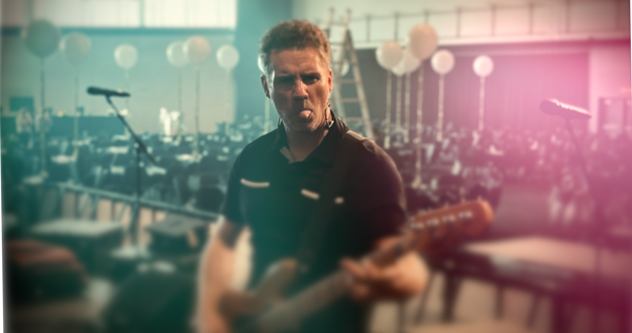 A man making a silly face with his tongue out, holding a guitar in a gym or studio with microphones, chairs, and exercise equipment in the background.