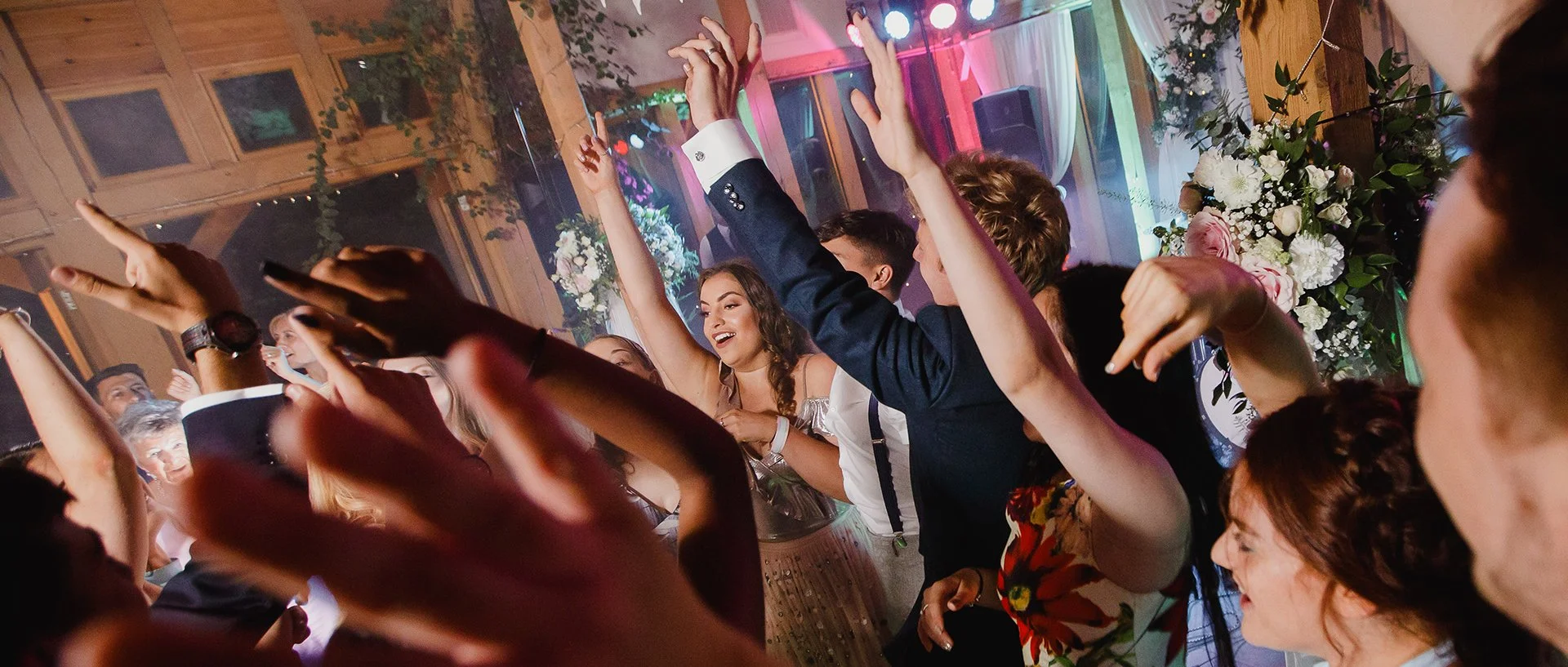People dancing at a celebration or party with colorful lights, floral decorations, and a lively atmosphere.
