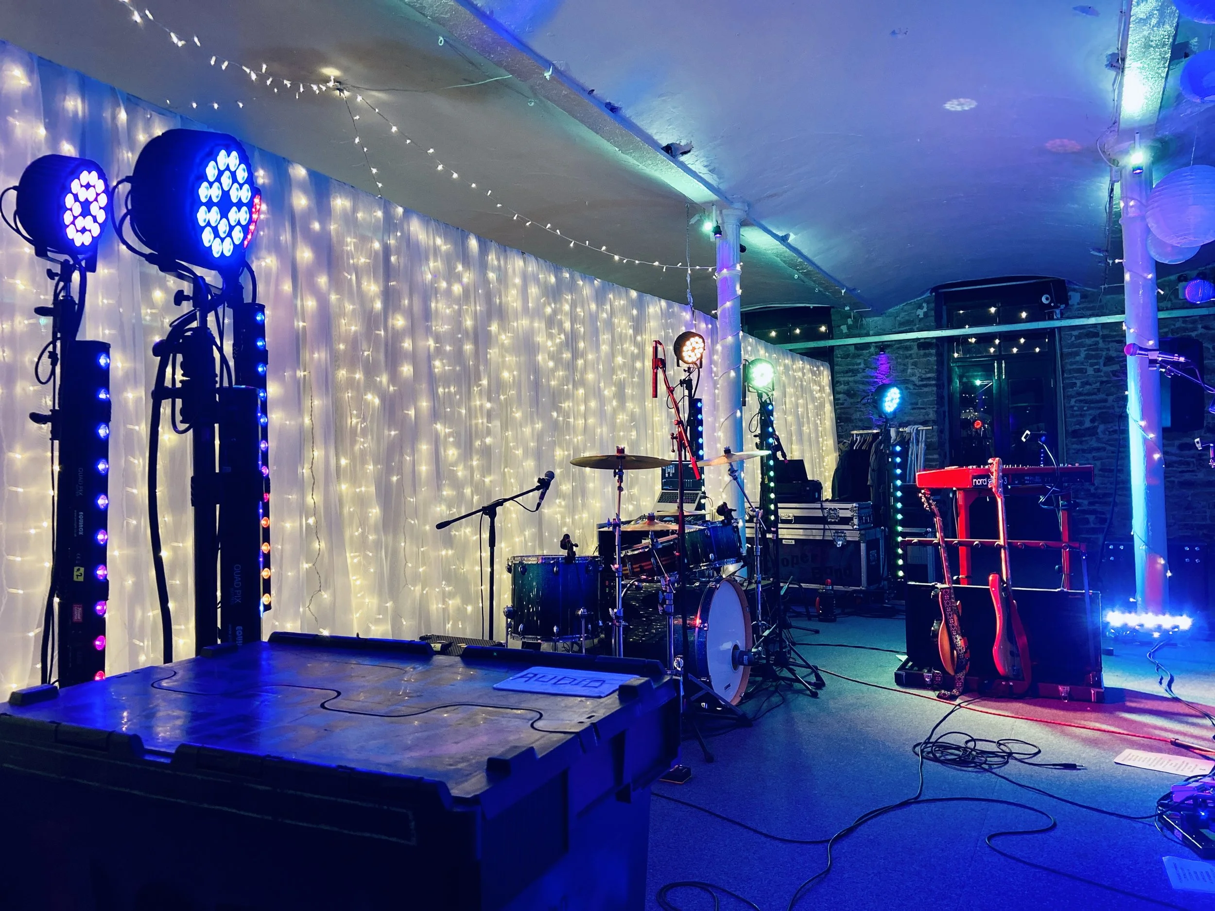 Music stage with instruments and colorful LED lighting, including drums, microphone, guitars, a keyboard, and stage lights, decorated with string lights against a backdrop with white curtains.
