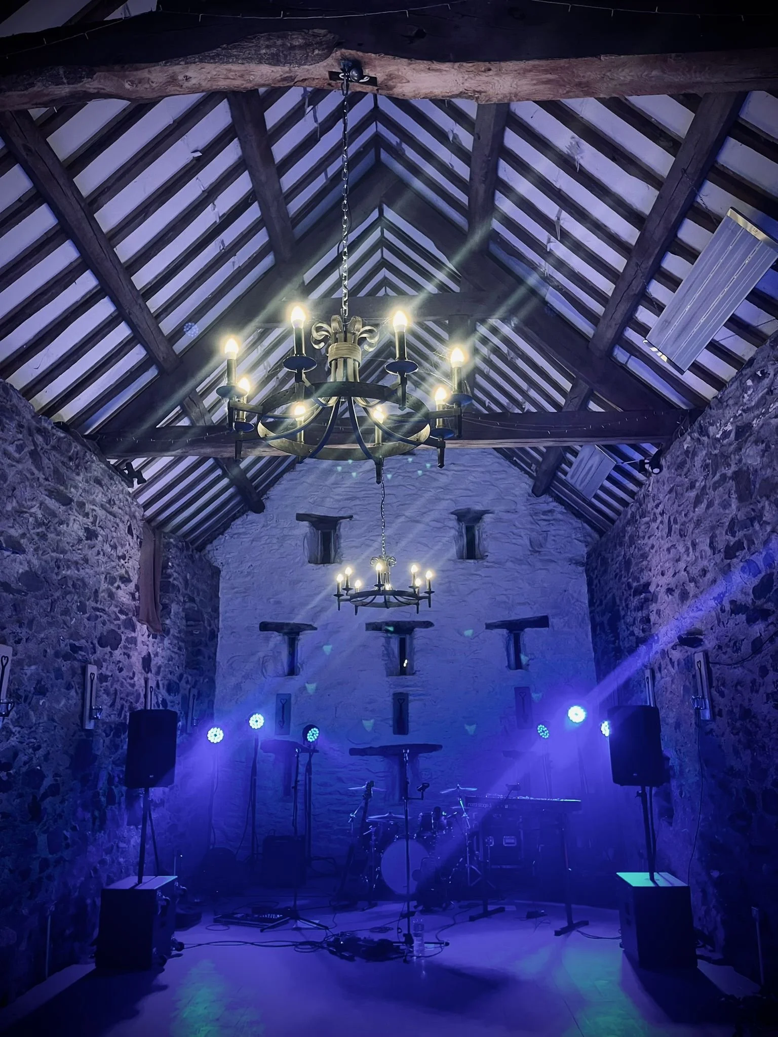 An indoor music venue with stone walls, wooden beams, and a high, vaulted ceiling. Two chandeliers hang from the ceiling, and stage lighting is directed toward a drum set and musical equipment at the back of the stage, with purple and blue lights cre