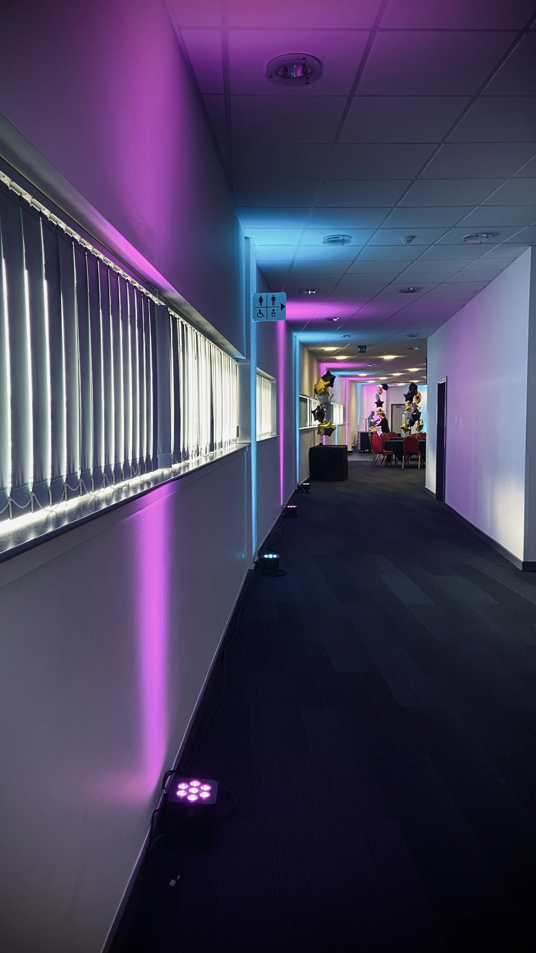 Hallway decorated with purple and blue ambient lighting leading to a social area with balloons and people seated at tables.