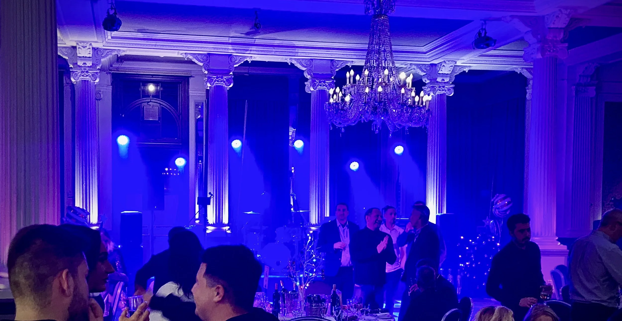 People socializing and dancing in an elegant banquet hall decorated with blue lighting, a large chandelier, and ornate white architectural details.