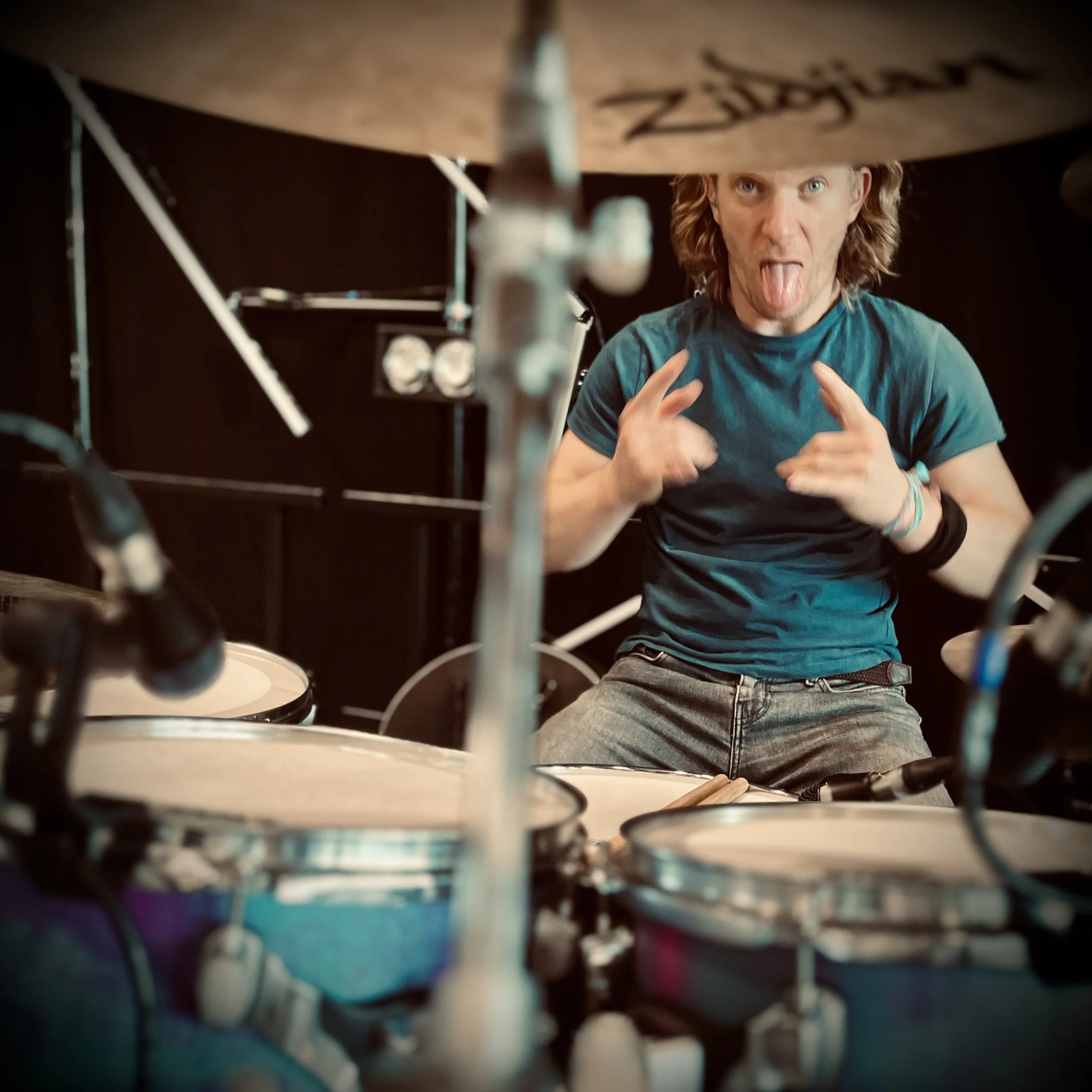 A man with long hair in a blue shirt sticking out his tongue and making a gesture, seen from behind the drum set he is playing.