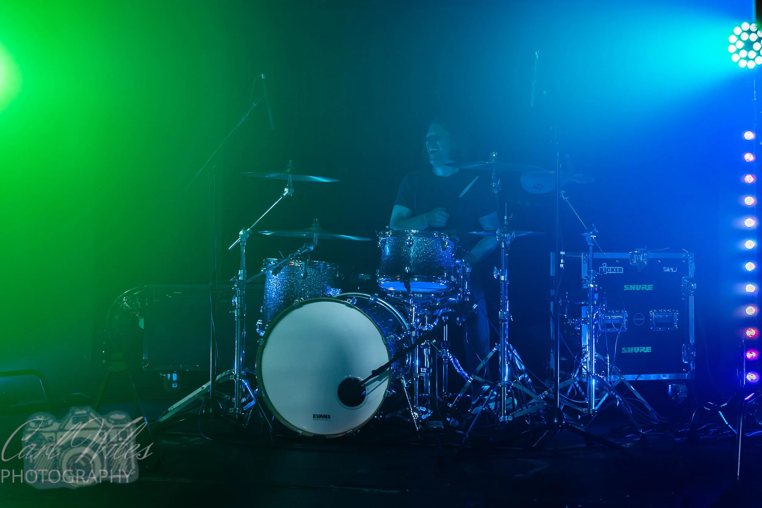 A drummer playing on stage with colorful green, blue, and purple stage lights illuminating the background.