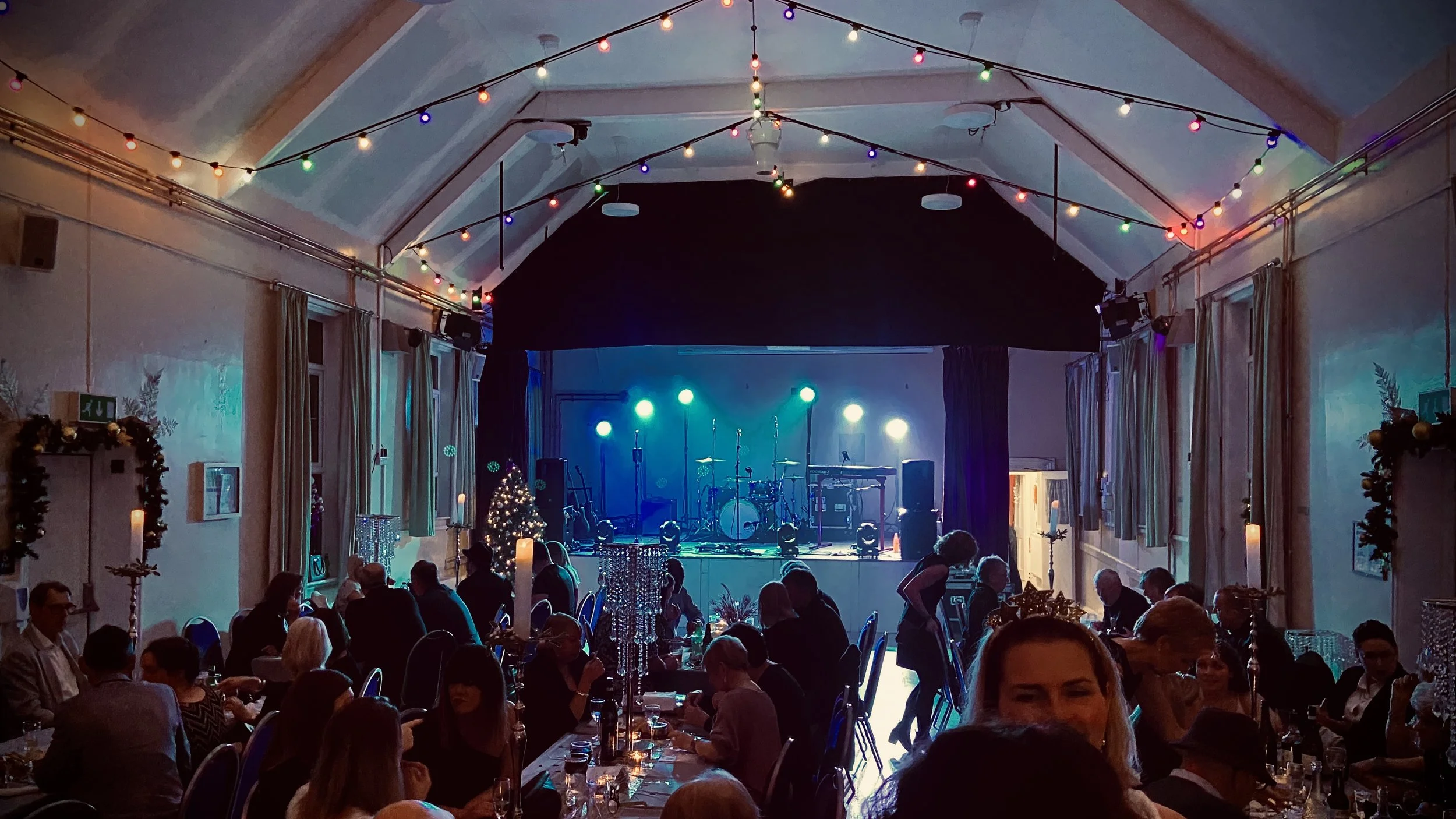 An indoor event space decorated for a celebration, with tables filled with people, a stage with musical instruments, Christmas tree, holiday decorations, and colorful string lights hanging from the ceiling.