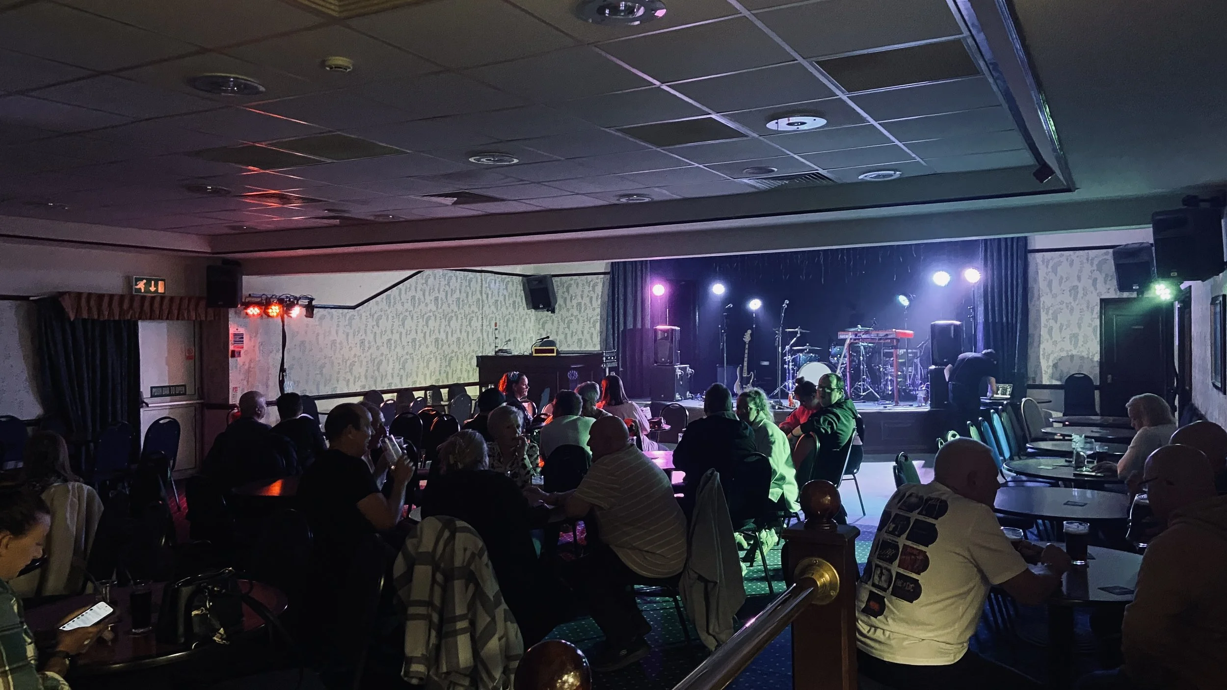 People seated at tables in a dimly lit venue with a stage set for live music, including instruments and microphones, illuminated by colorful stage lights.