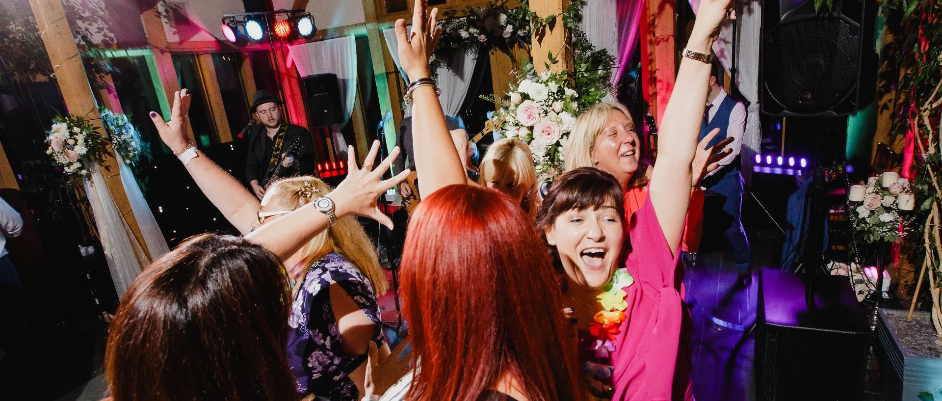 People enjoying and dancing at a lively celebration or party with a band playing in the background and floral decorations.