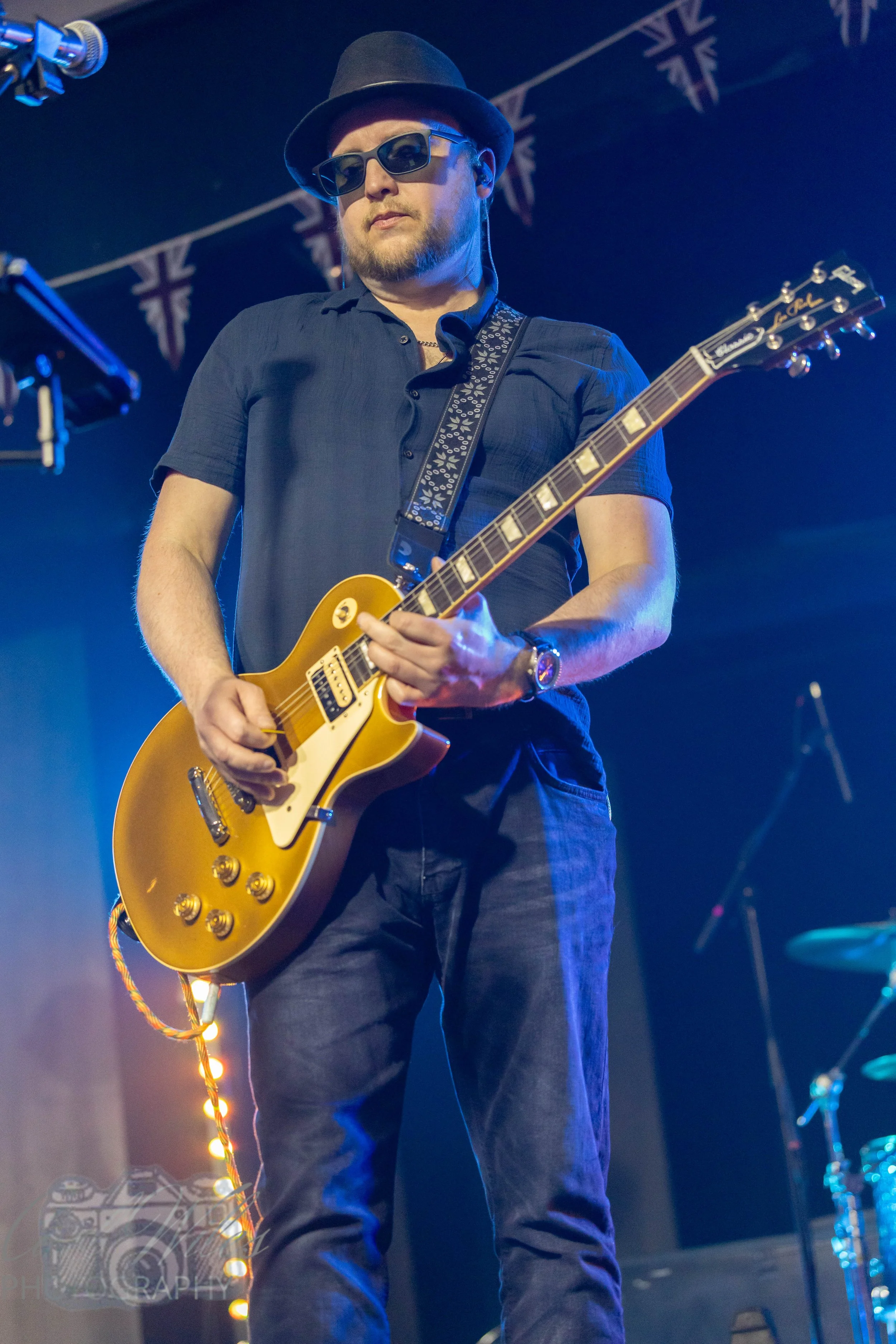 A man wearing sunglasses, a fedora, a short-sleeved shirt, and jeans playing a yellow electric guitar on stage with blue lighting and a backdrop with Union Jack flags.