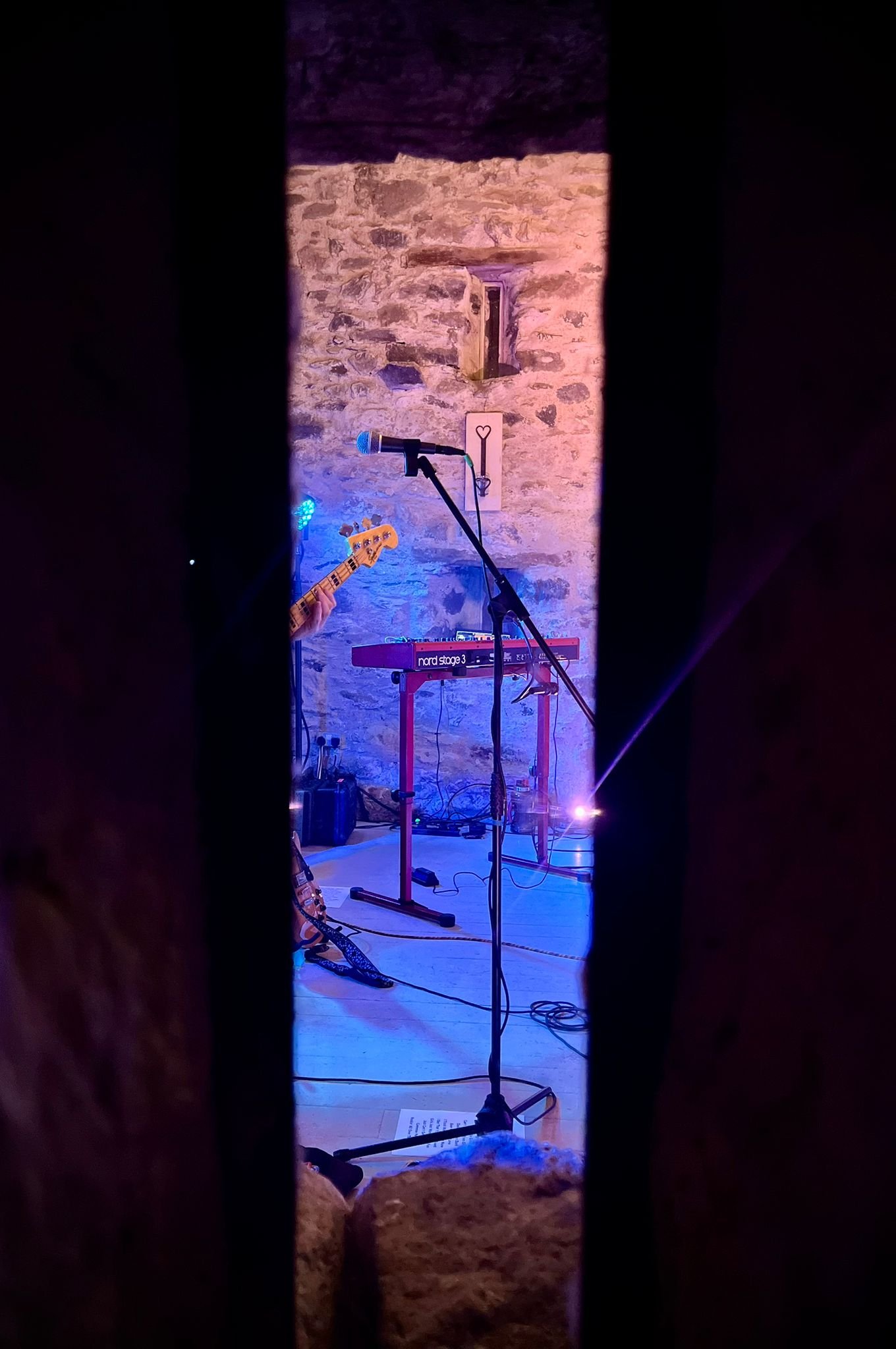 View of a stage with musical instruments, including a keyboard on a stand, microphone, and guitar, set against a stone wall, seen through a narrow opening.