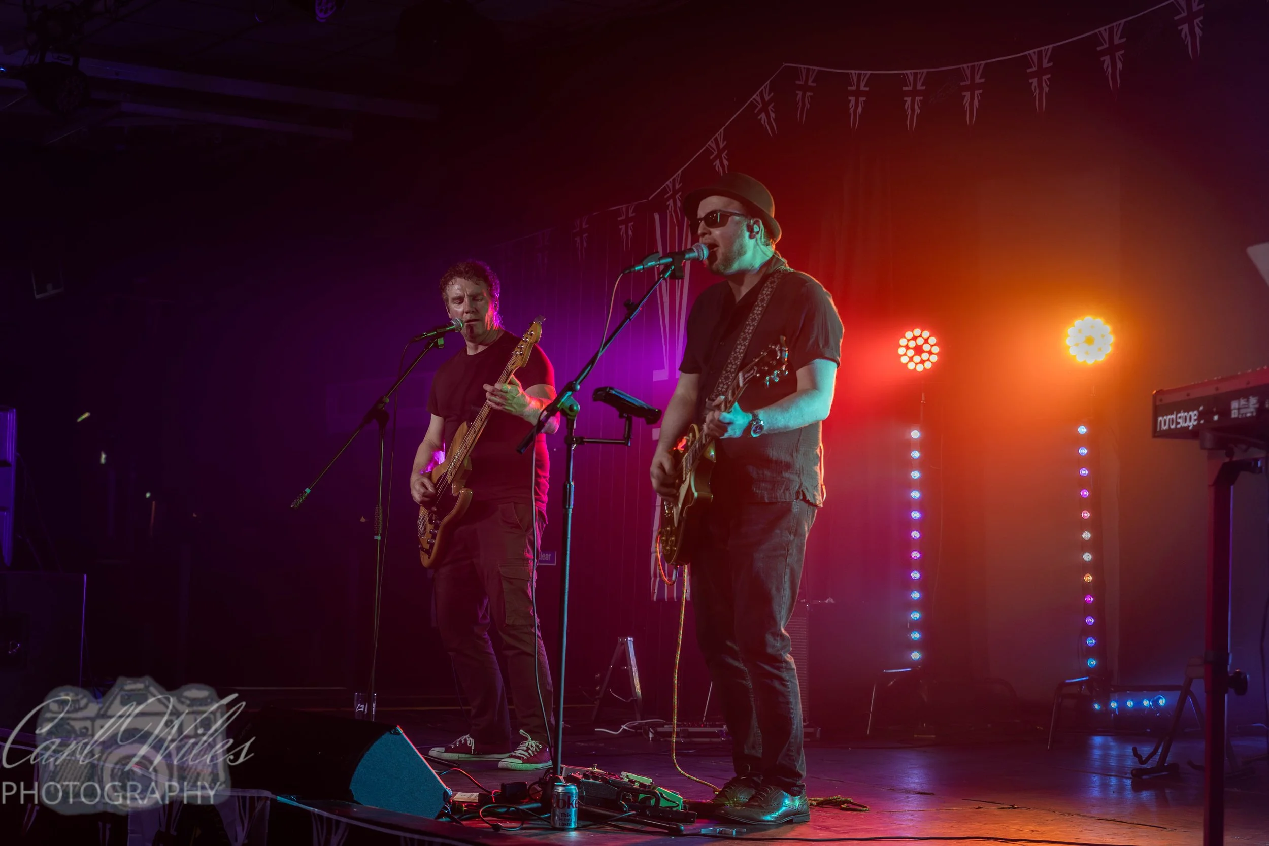 Two musicians performing on stage with colorful lighting and a string of British flags hanging overhead.