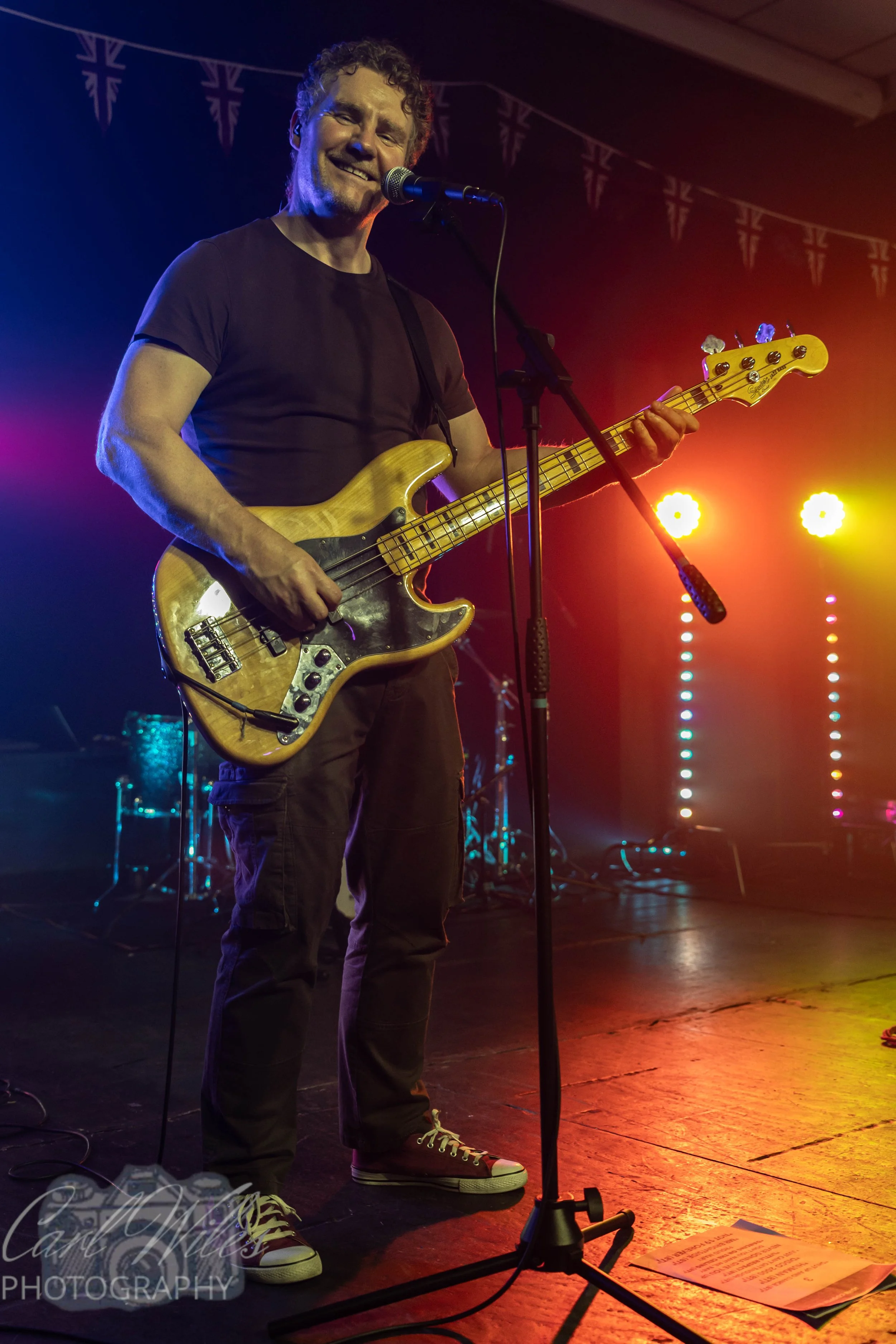 A man performing on stage with a bass guitar, smiling in front of colorful stage lights, with a drum set in the background.