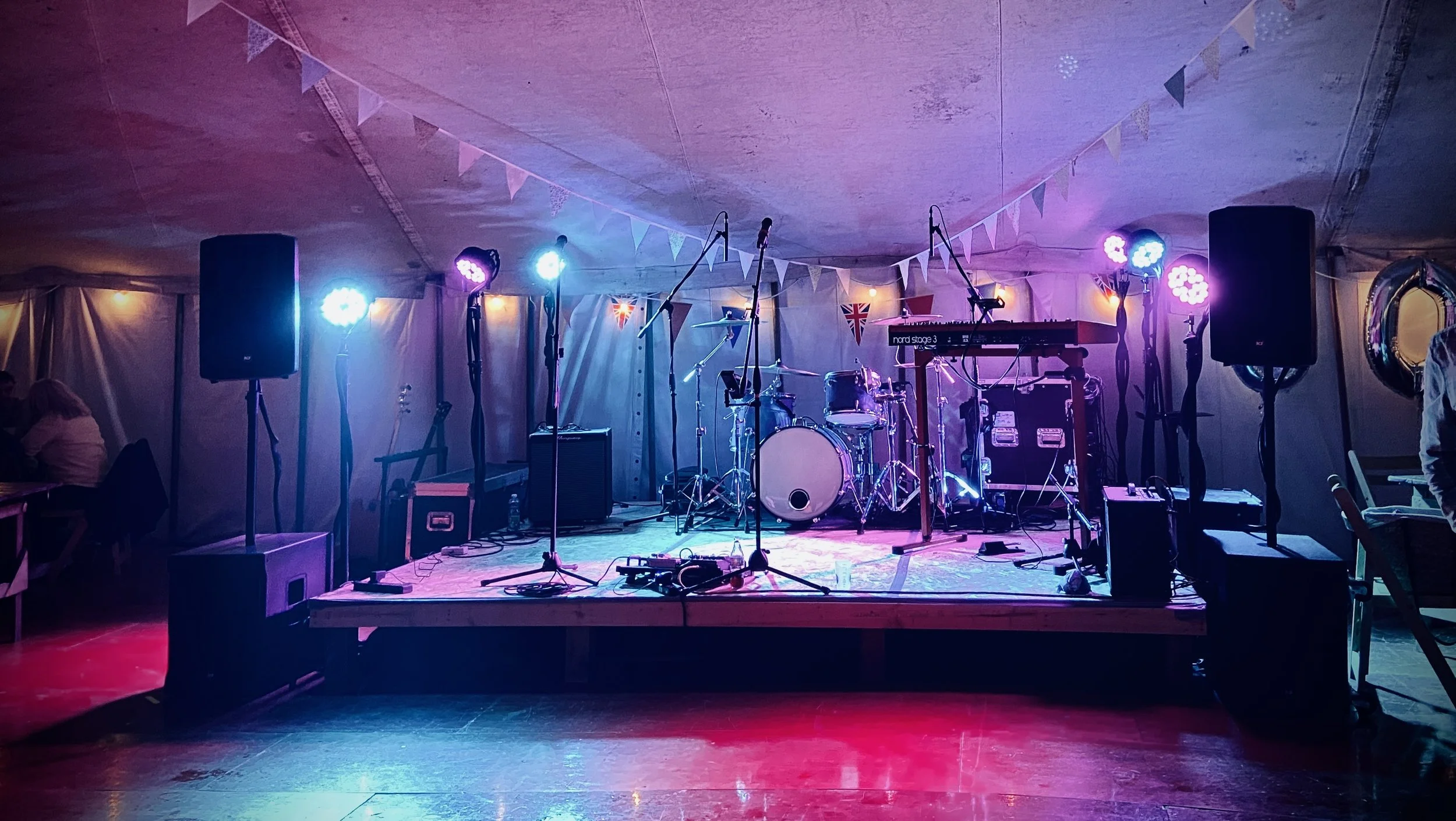 Stage set up with drum kit, keyboard, microphone stands, speakers, and stage lights in a tent decorated with bunting flags