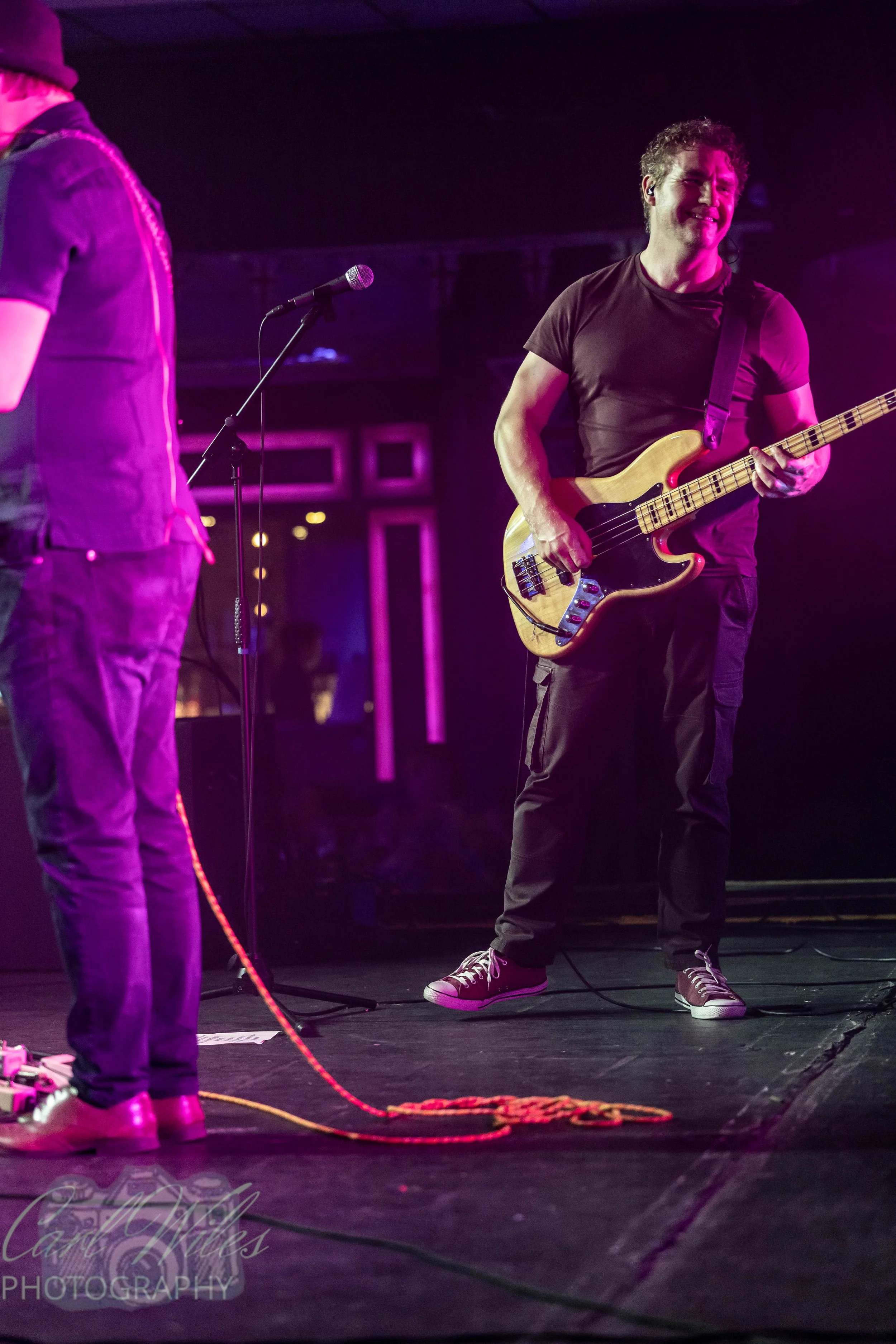 Close-up of a musician playing an electric bass guitar on stage with purple lighting. The musician is wearing a black t-shirt, black pants, and sneakers, with headphones. Part of another person wearing a denim vest and camouflage pants is visible on 
