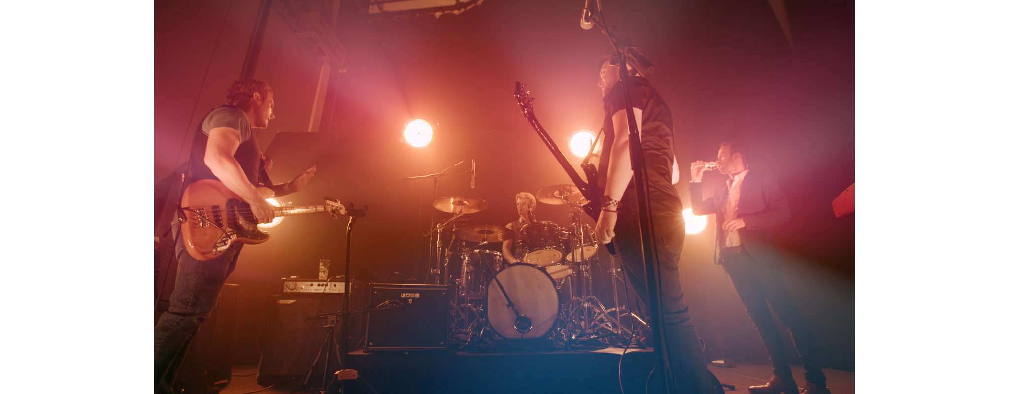 A live band performing on stage illuminated by bright pink and orange lights, featuring a guitarist, a drummer, a vocalist, and another band member.