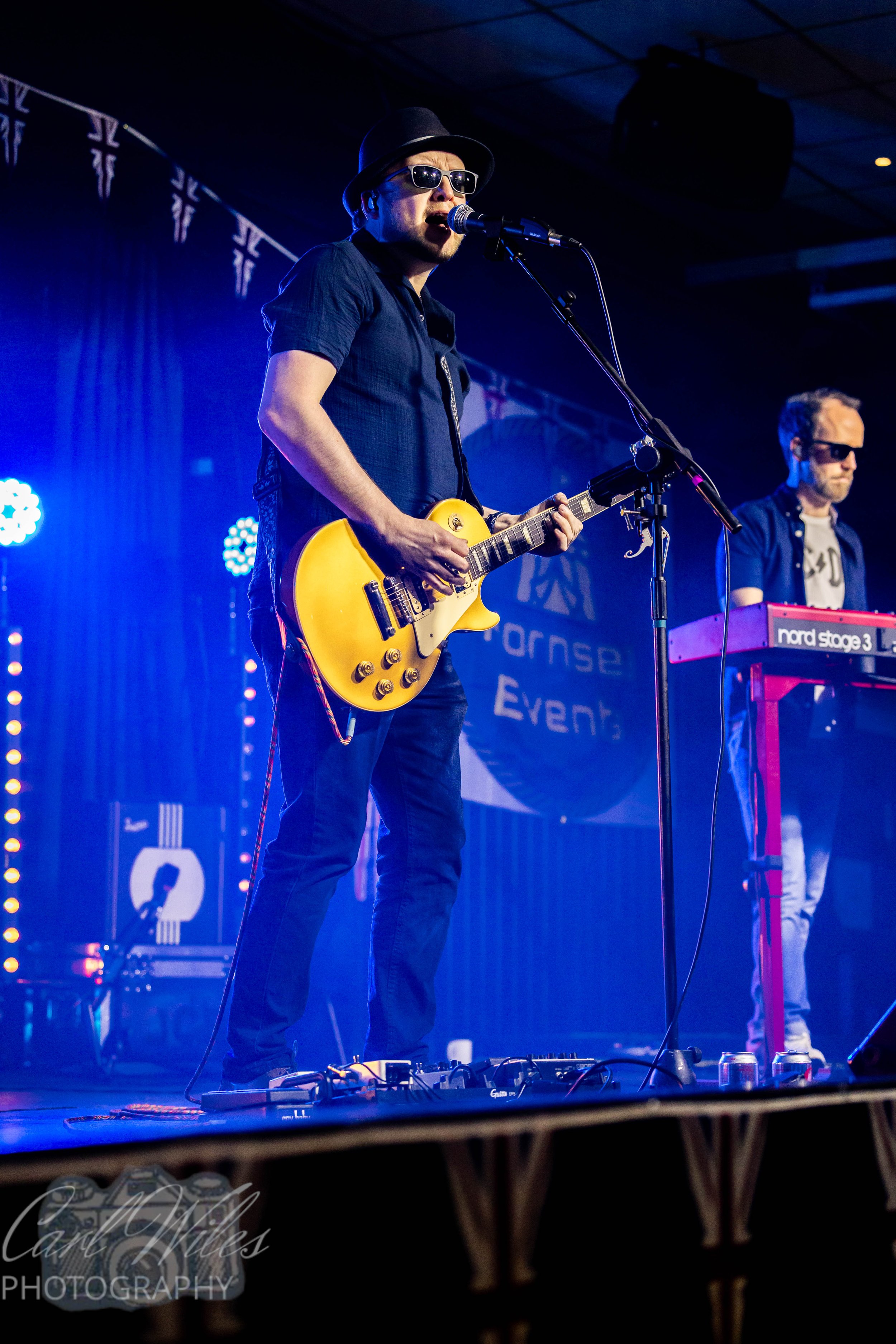 Male musician wearing sunglasses, a black hat, and a dark shirt, playing an electric guitar and singing into a microphone on stage with blue lighting.