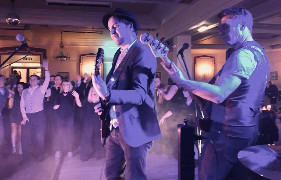 Two musicians playing electric guitars on stage at a lively indoor event with an audience dancing and enjoying the music.