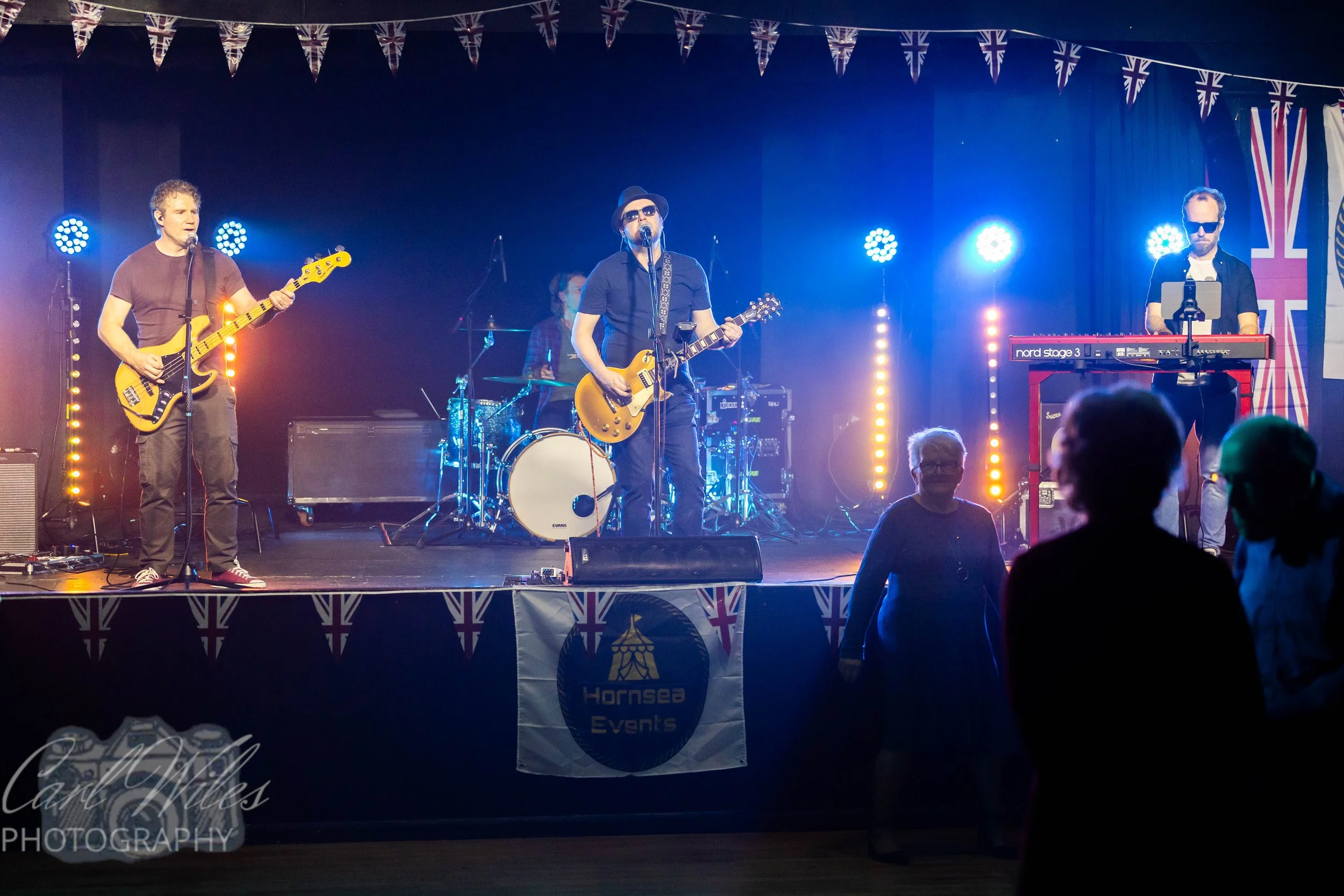 A band performing on stage at an event decorated with Union Jack flags. The band consists of a guitarist, a drummer in the background, and a keyboardist to the right. Audience members are visible in the foreground.