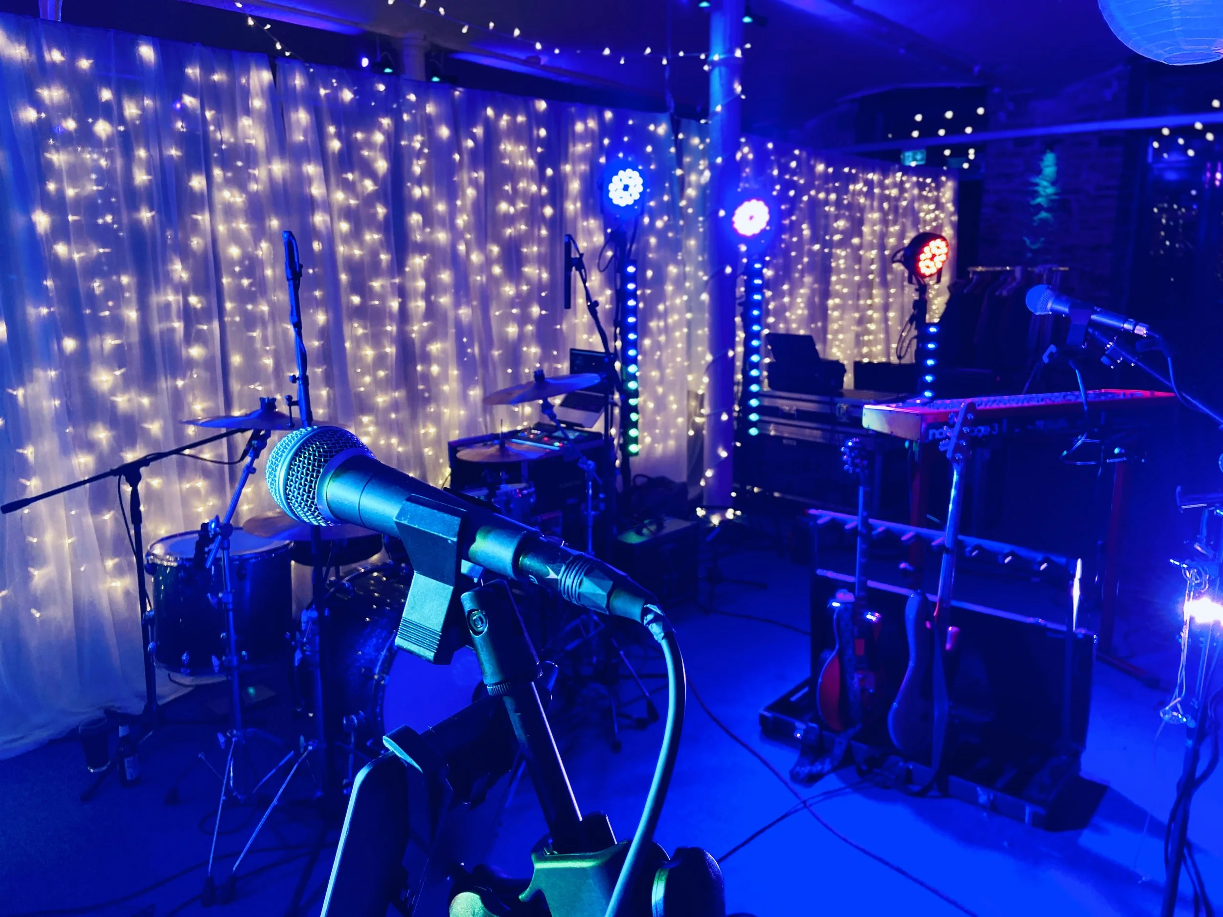 Music performance stage with microphone, drums, guitars, and keyboard, illuminated by colorful stage lights and string lights in the background.