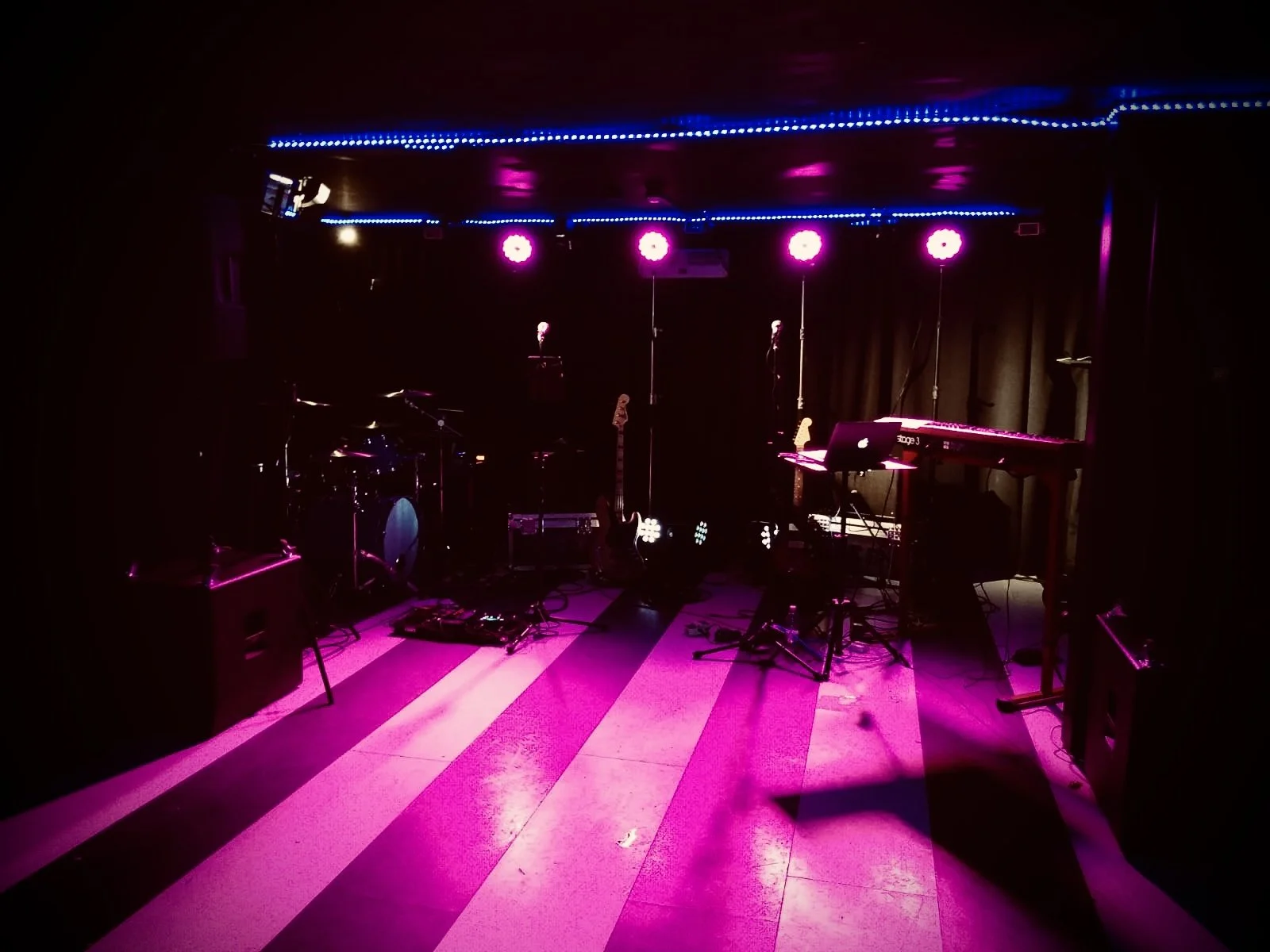 Stage with musical instruments and pink and purple lighting, including drums, electric guitars, keyboards, microphones, and other equipment.