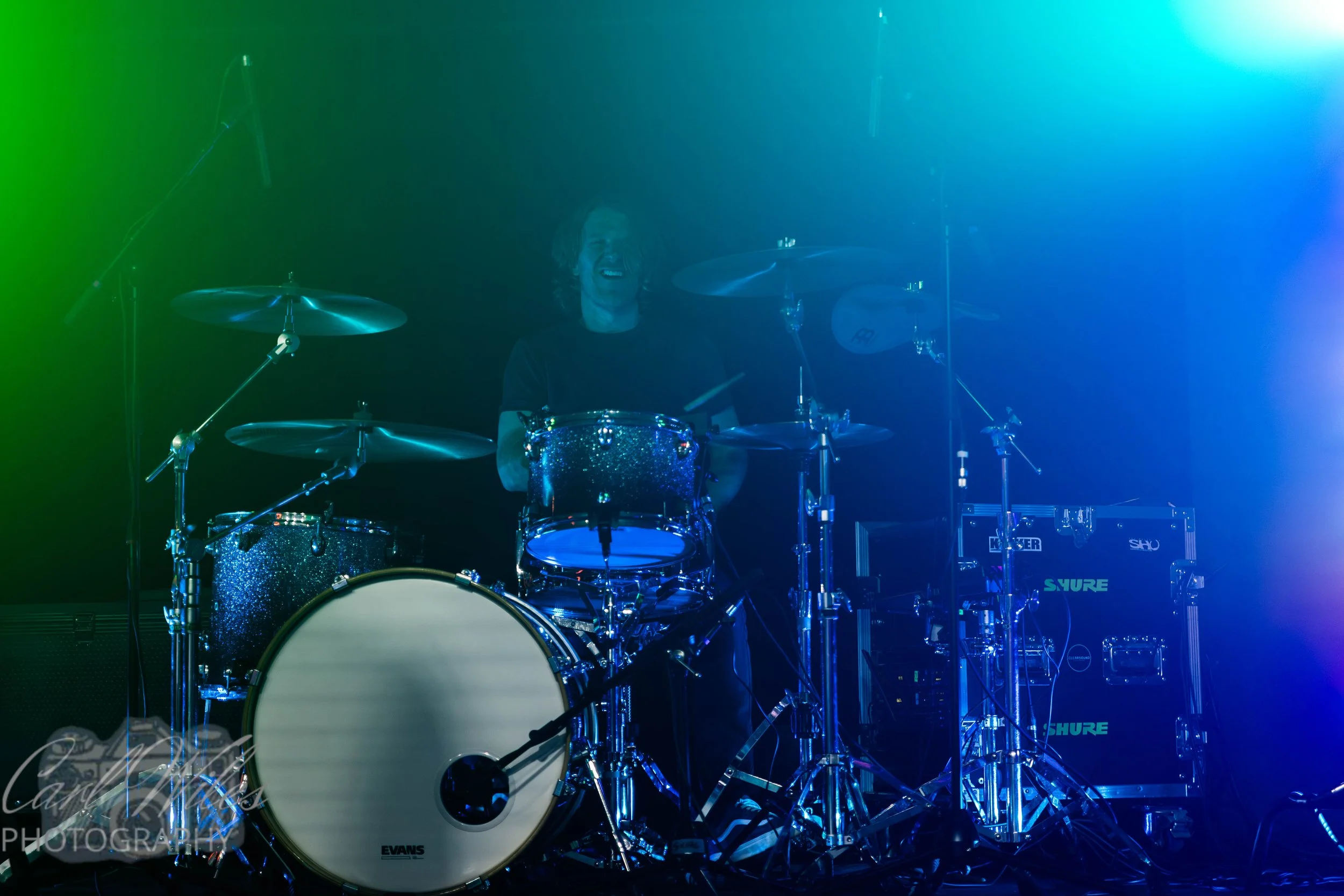A person playing a drum set on stage surrounded by blue and green stage lighting.