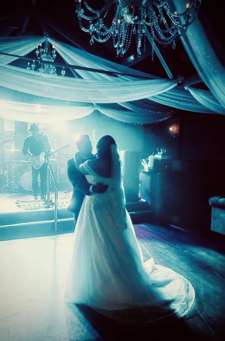 A bride and groom dance together under decorative drapery and a chandelier at a wedding reception. Live music is played by a band in the background.