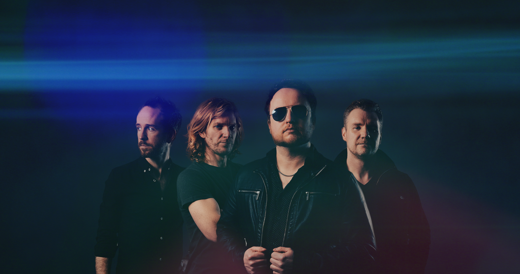 Four men standing against a dark background with colorful light streaks across the top, dressed in dark clothing, with one wearing sunglasses.