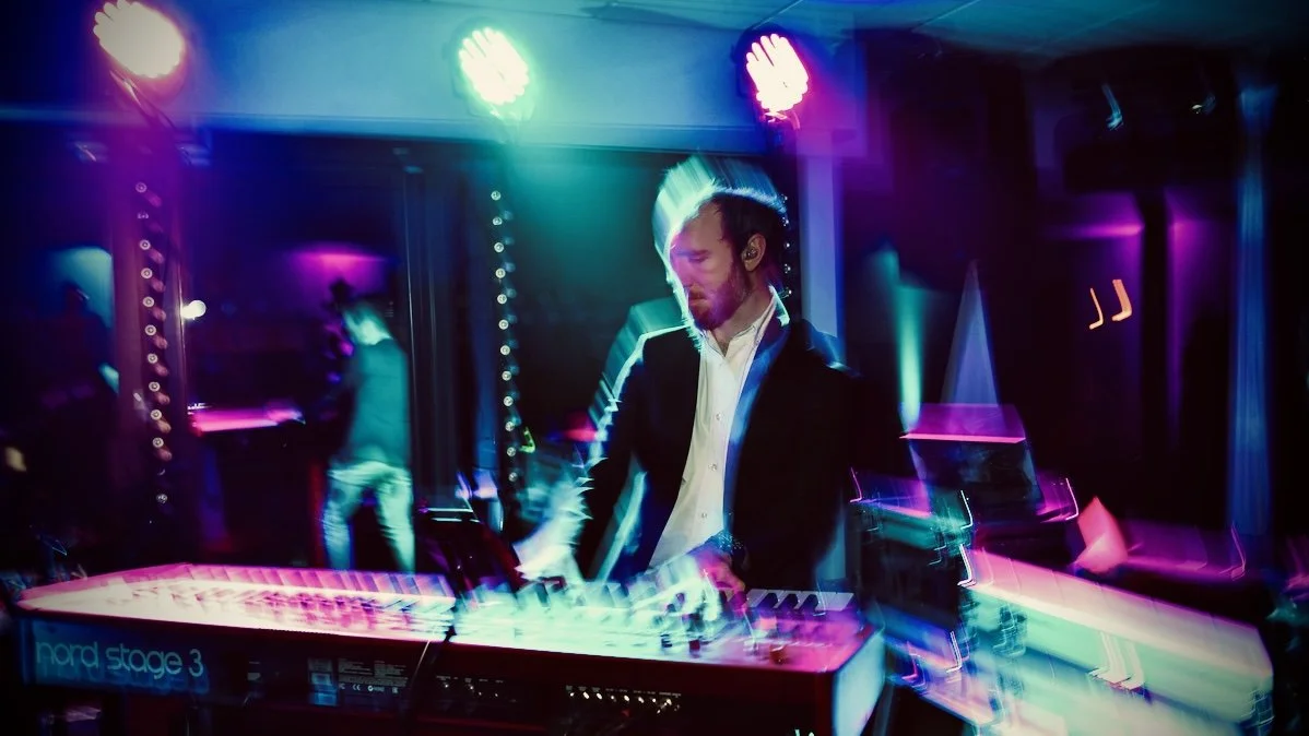 A man playing a keyboard on stage with colorful lights and a blurred background at a live music venue.