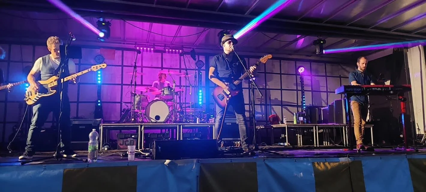 A band performing live on stage with four members playing instruments, colorful stage lighting, and a dark background.