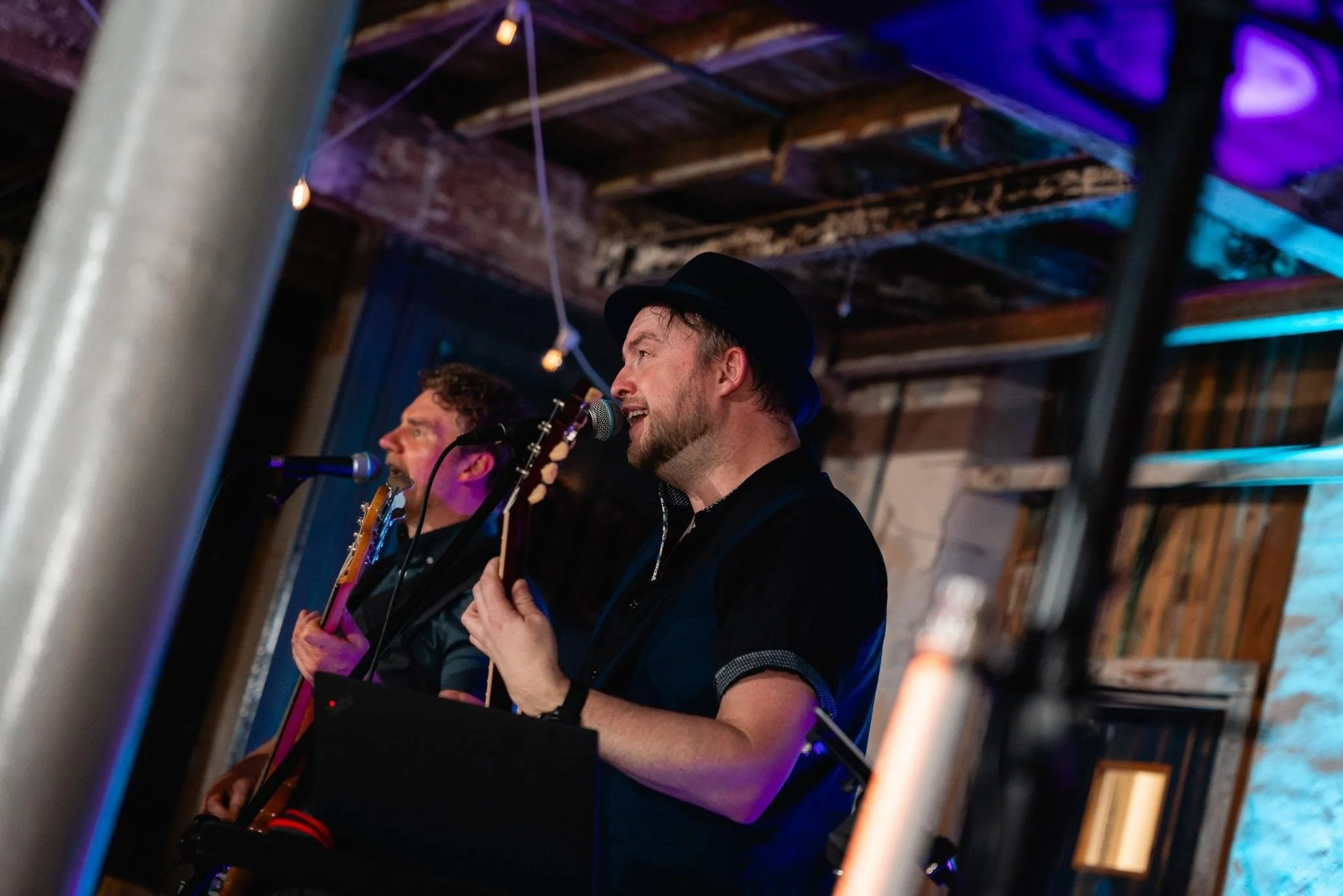 Two male musicians singing and playing guitars on stage inside a rustic venue with wooden beams, string lights, and vibrant blue and purple lighting.