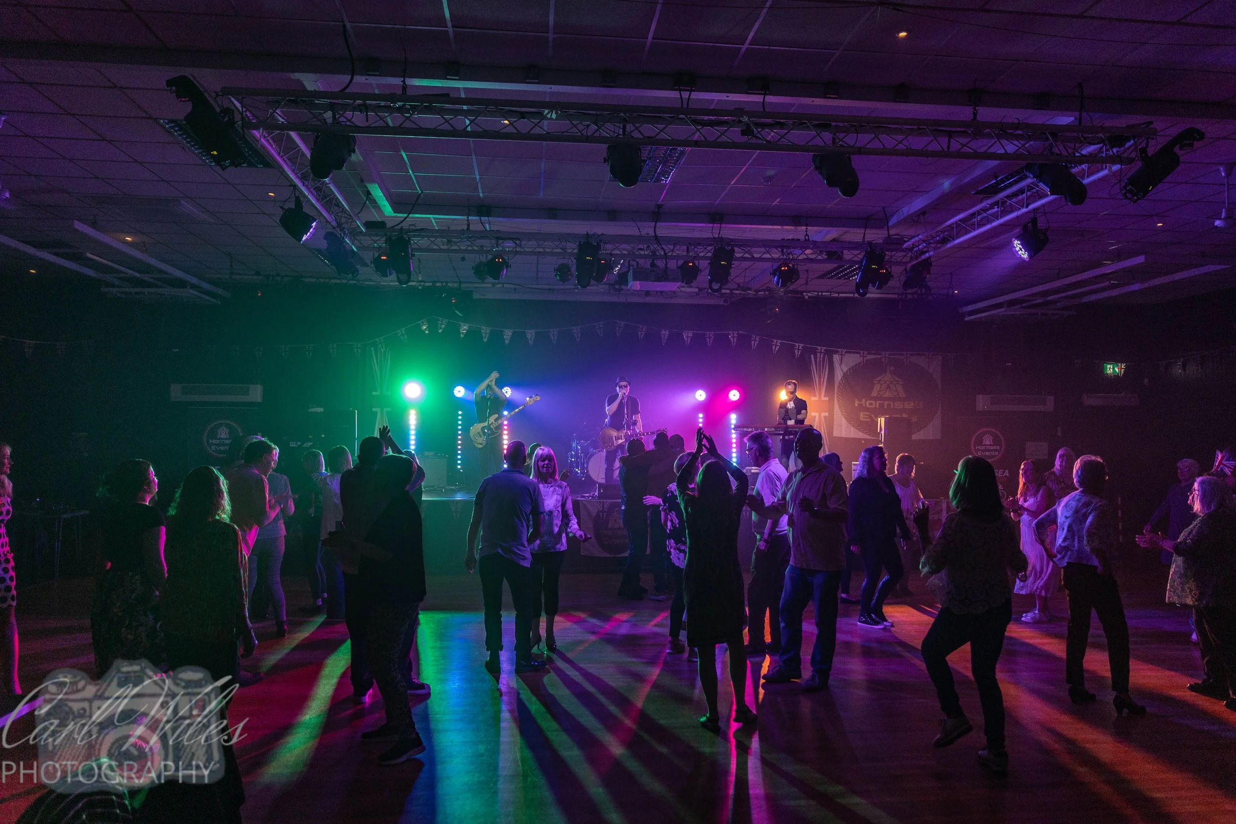 People dancing and enjoying a live music performance on stage in a dimly lit venue with colorful stage lighting.